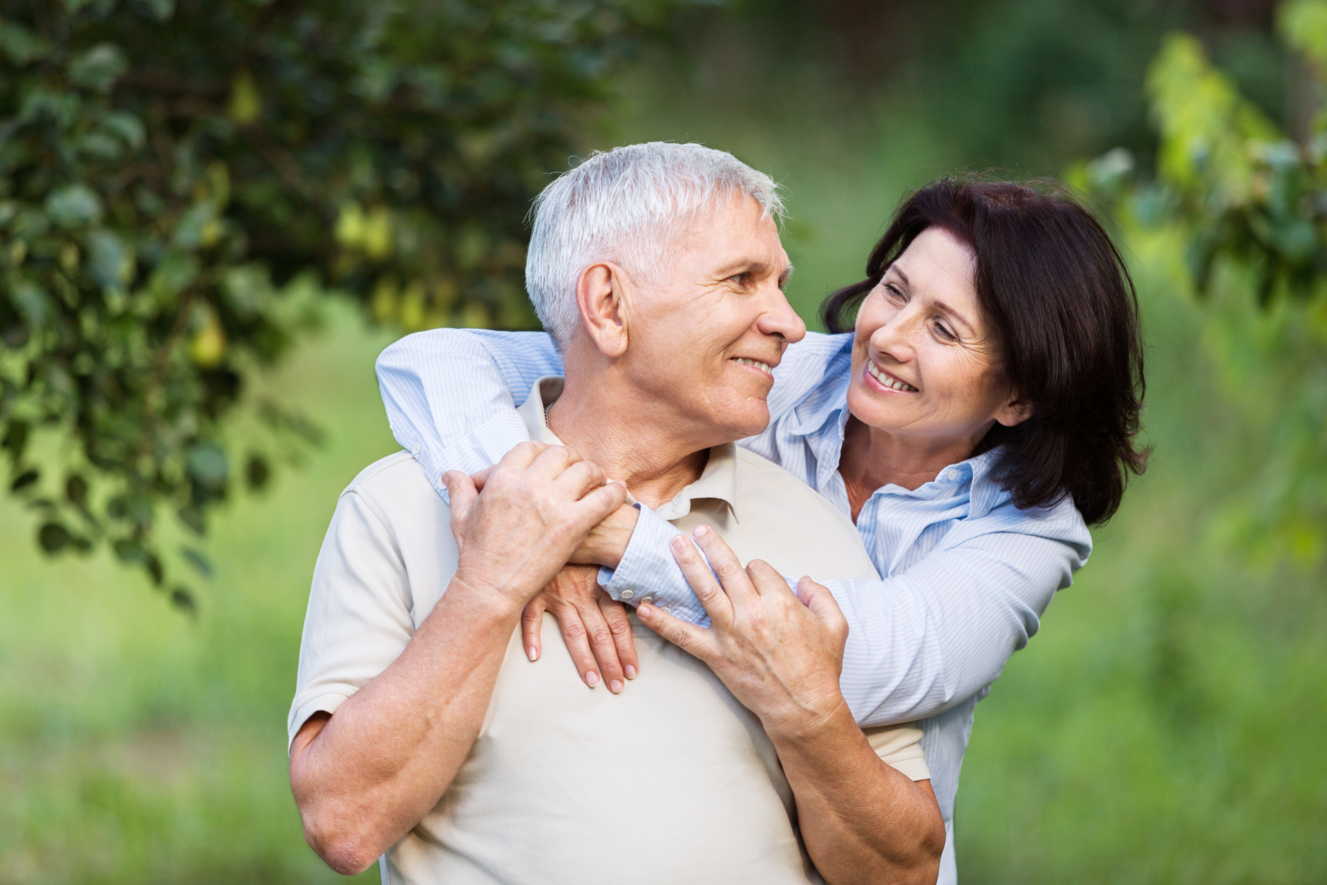 An older couple in a park, the woman has her arms around the man. They both smile, with greenery in the background.