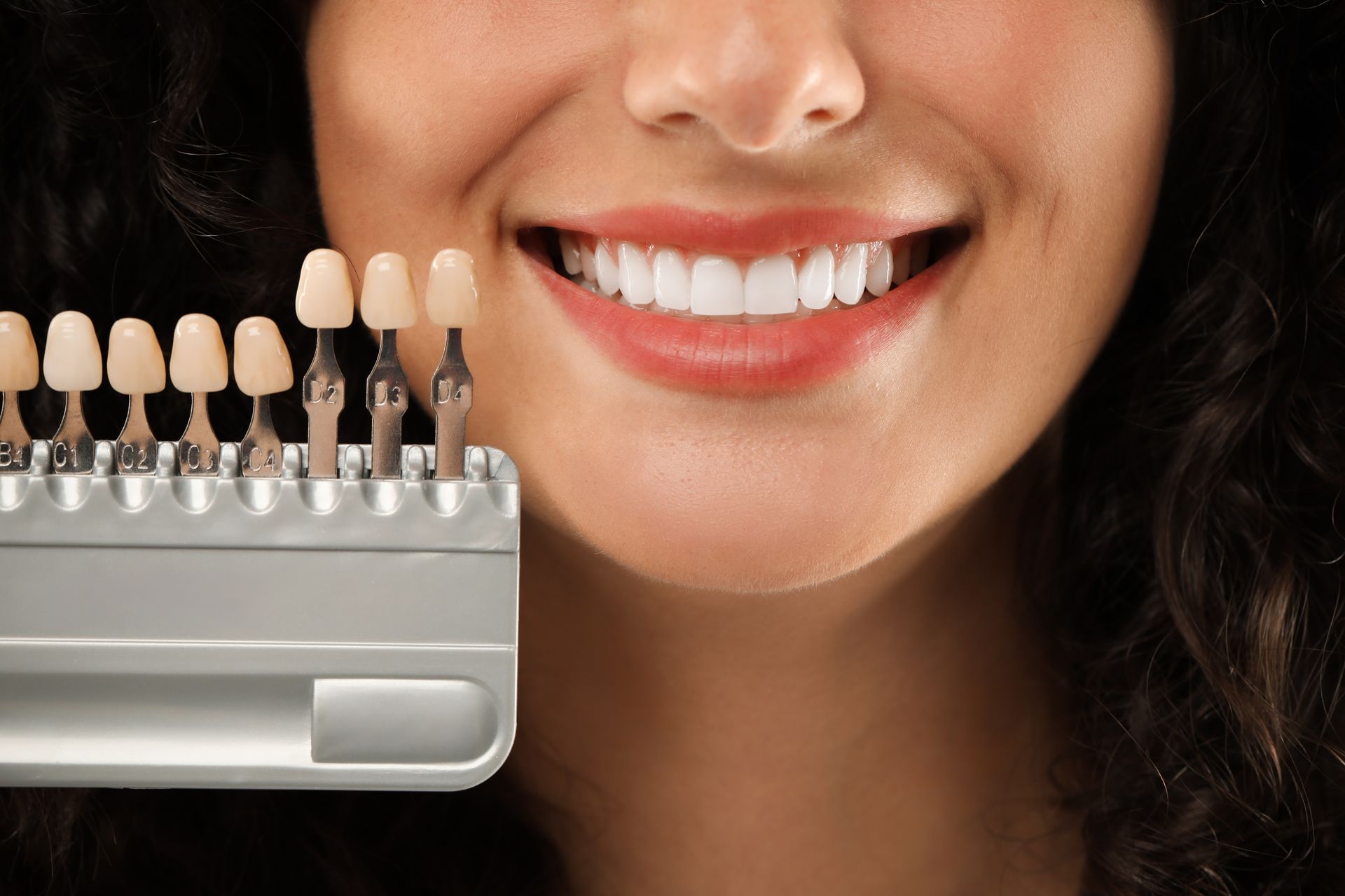 Woman smiling with bright white teeth next to a dental shade guide, showing tooth color options.