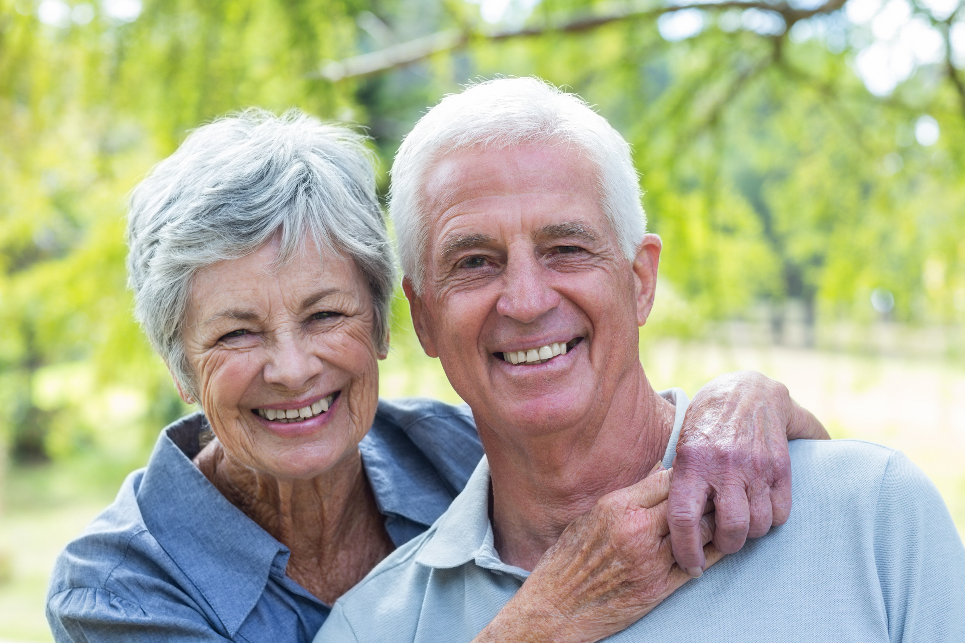 Elderly couple smiling at the camera outdoors, woman with arm around man's shoulders. Green background.
