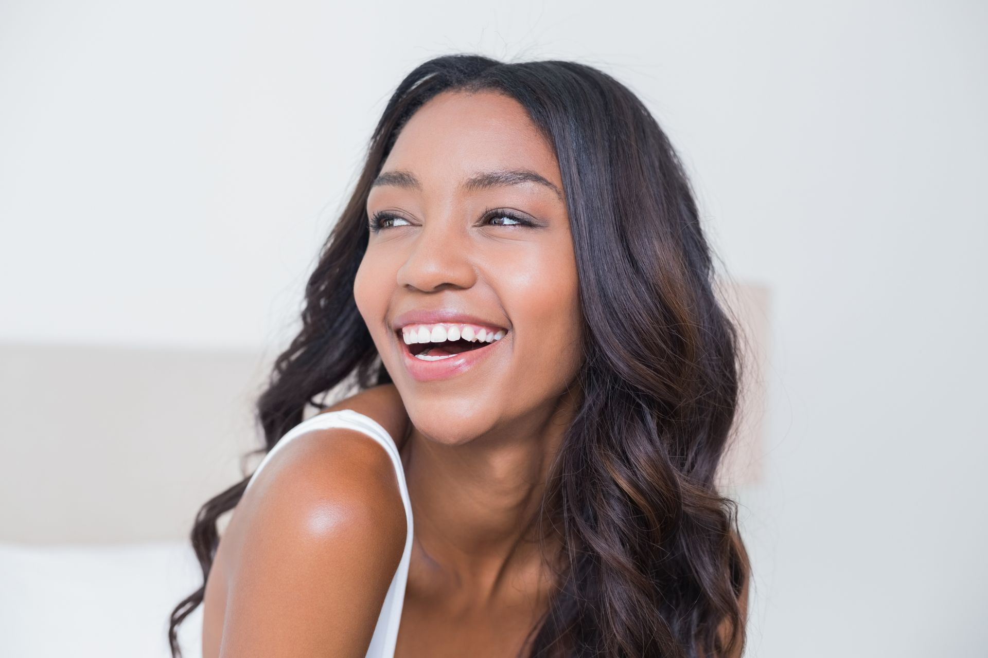Smiling Black woman with long, wavy dark hair, sitting on a bed in a bright room.
