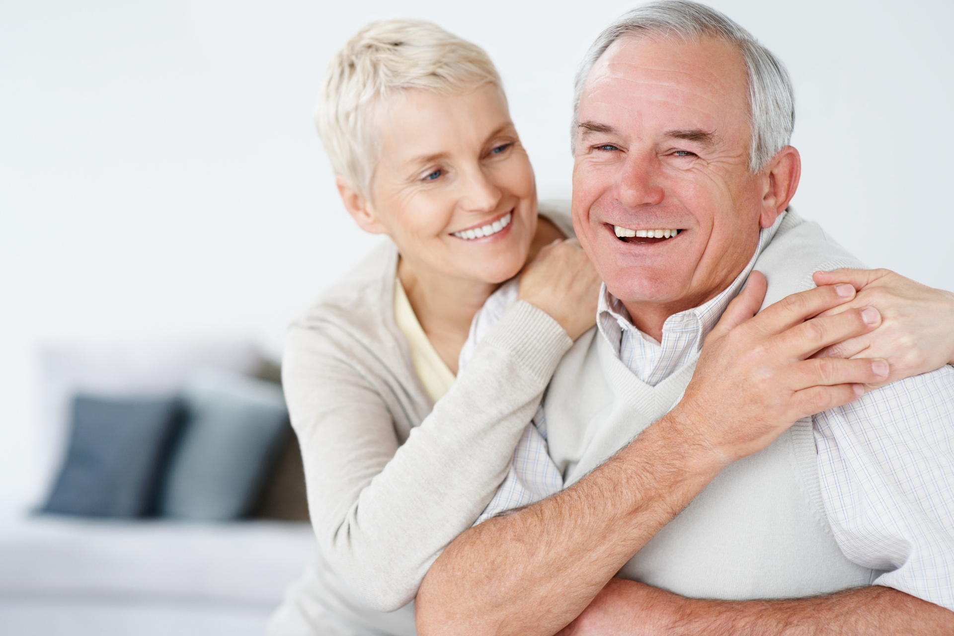 Smiling older couple embracing, woman with blonde hair has her arms around the man's shoulders. They are indoors, likely in a home.