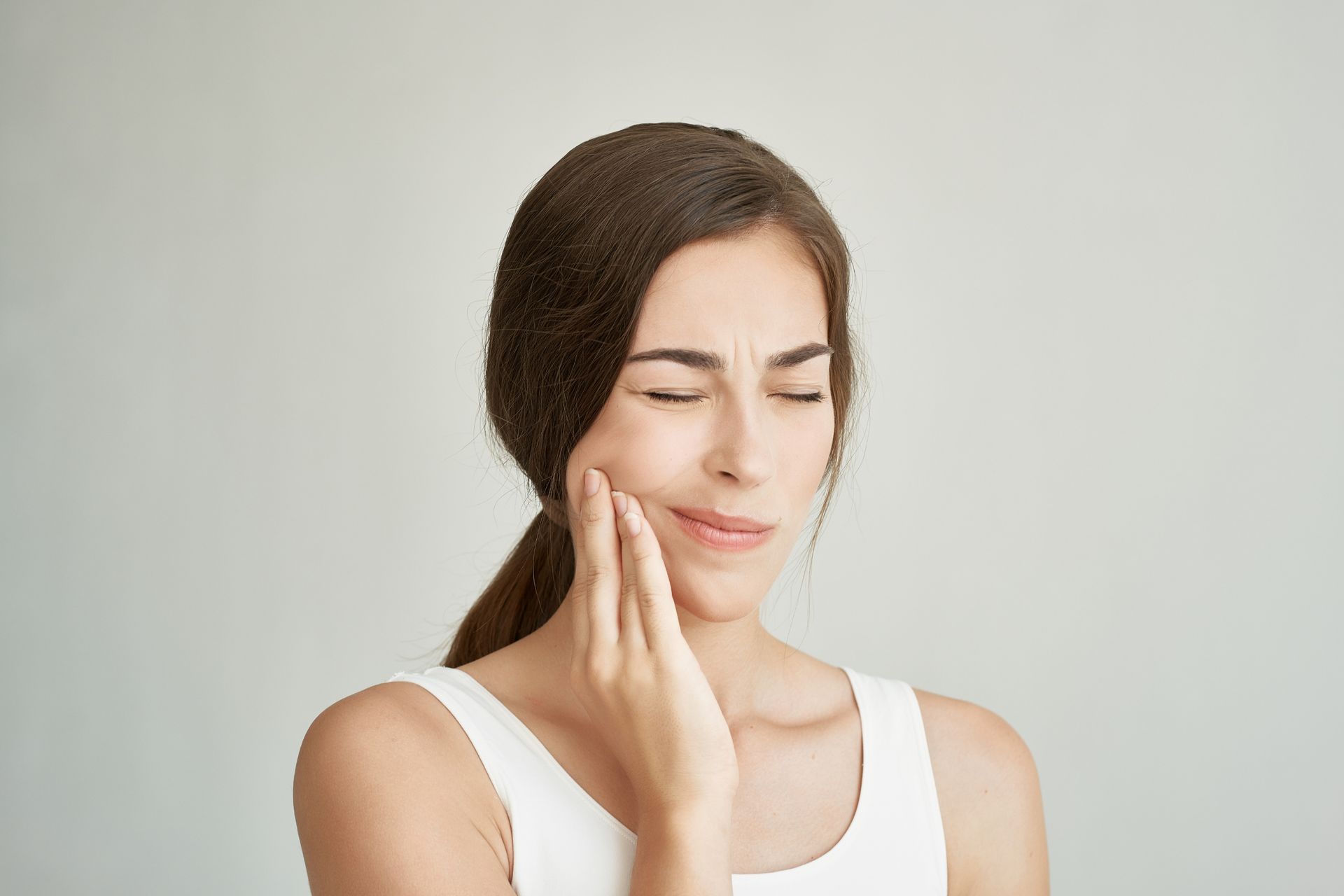 Woman with brown hair, wearing a white tank top, holds her hand to her cheek, indicating pain. Her eyes are closed.