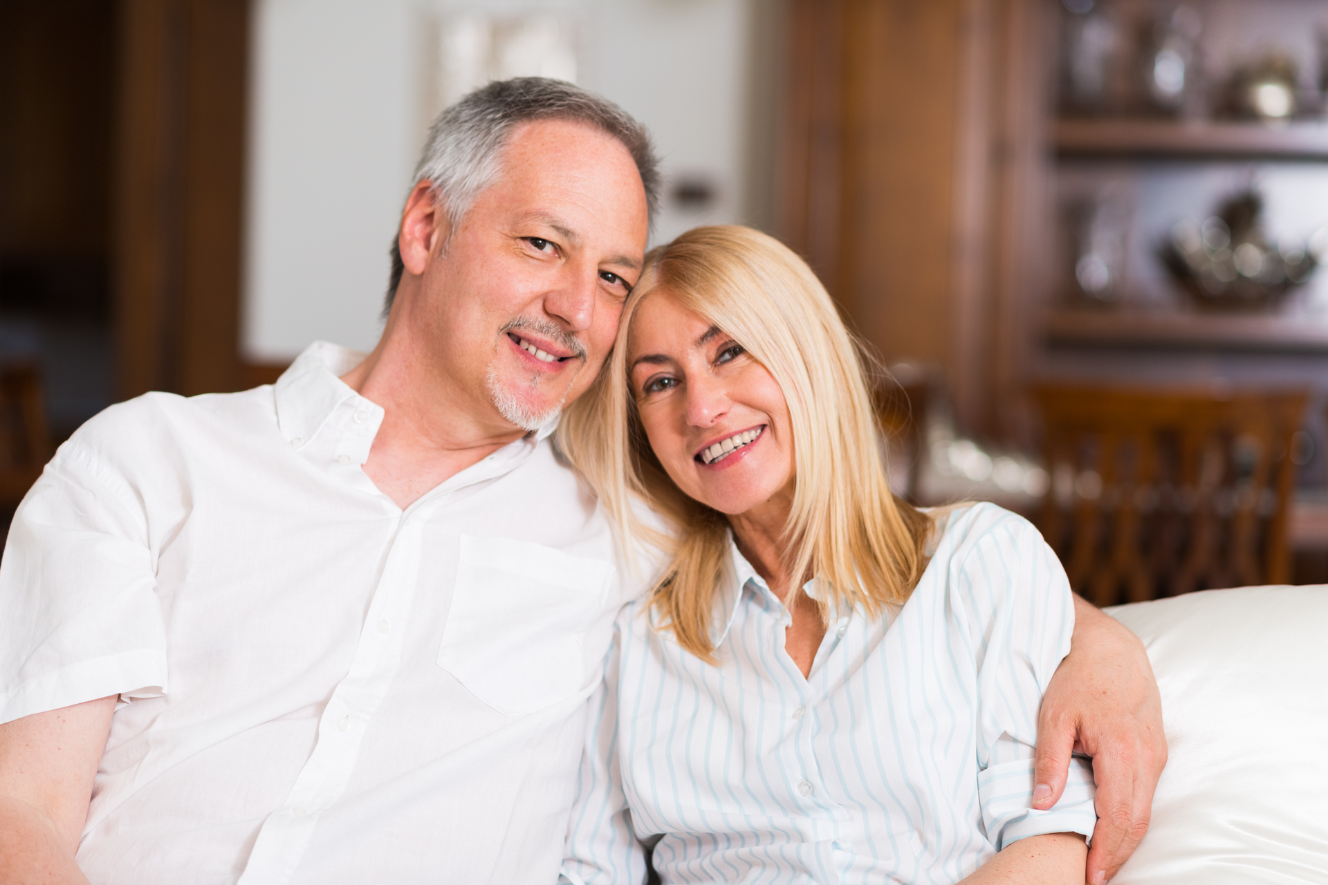 Smiling older couple sitting closely together on a sofa, man with arm around woman, home interior.
