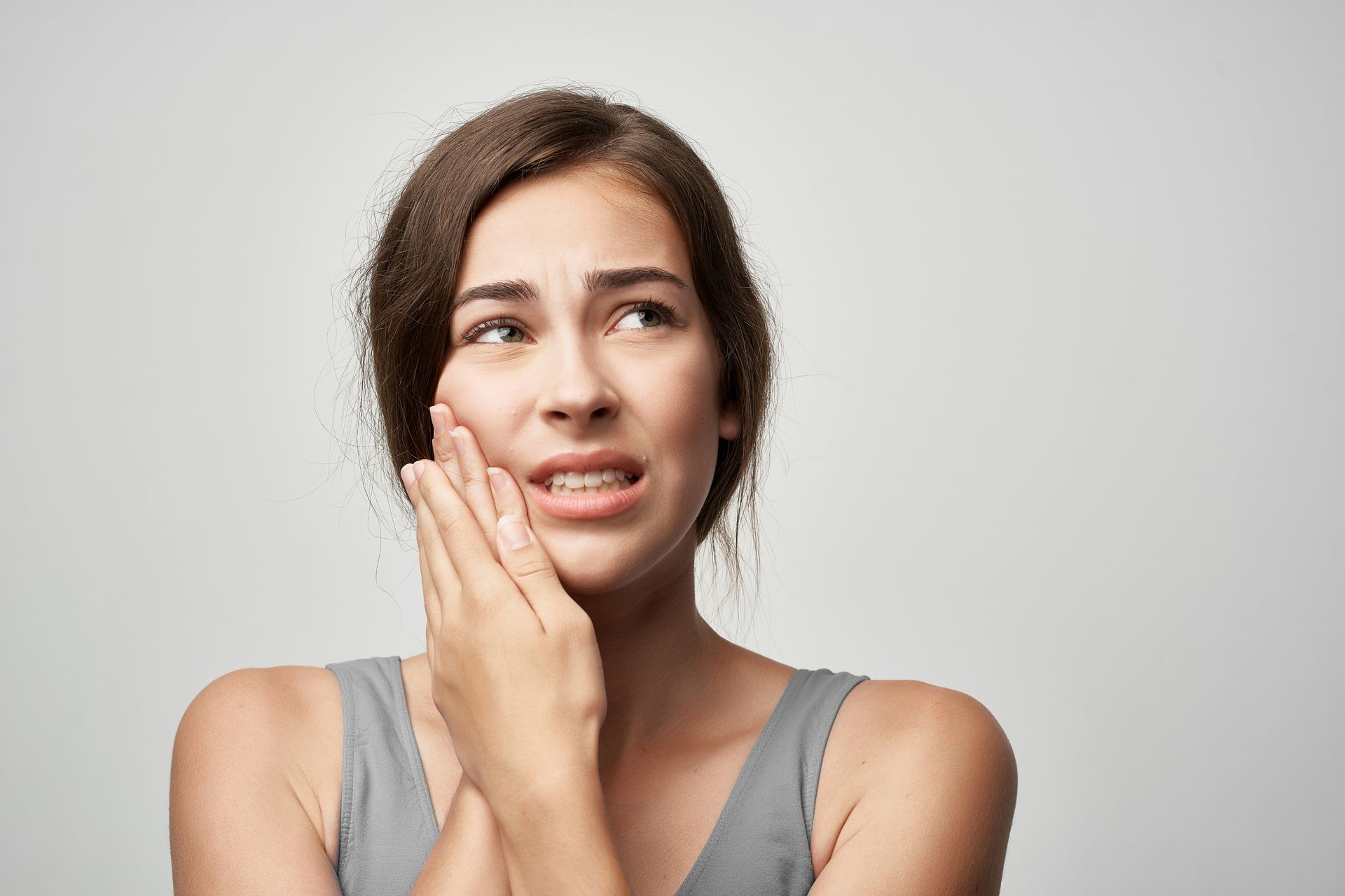 Woman in a gray tank top with an expression of pain, clutching her jaw with her hand.