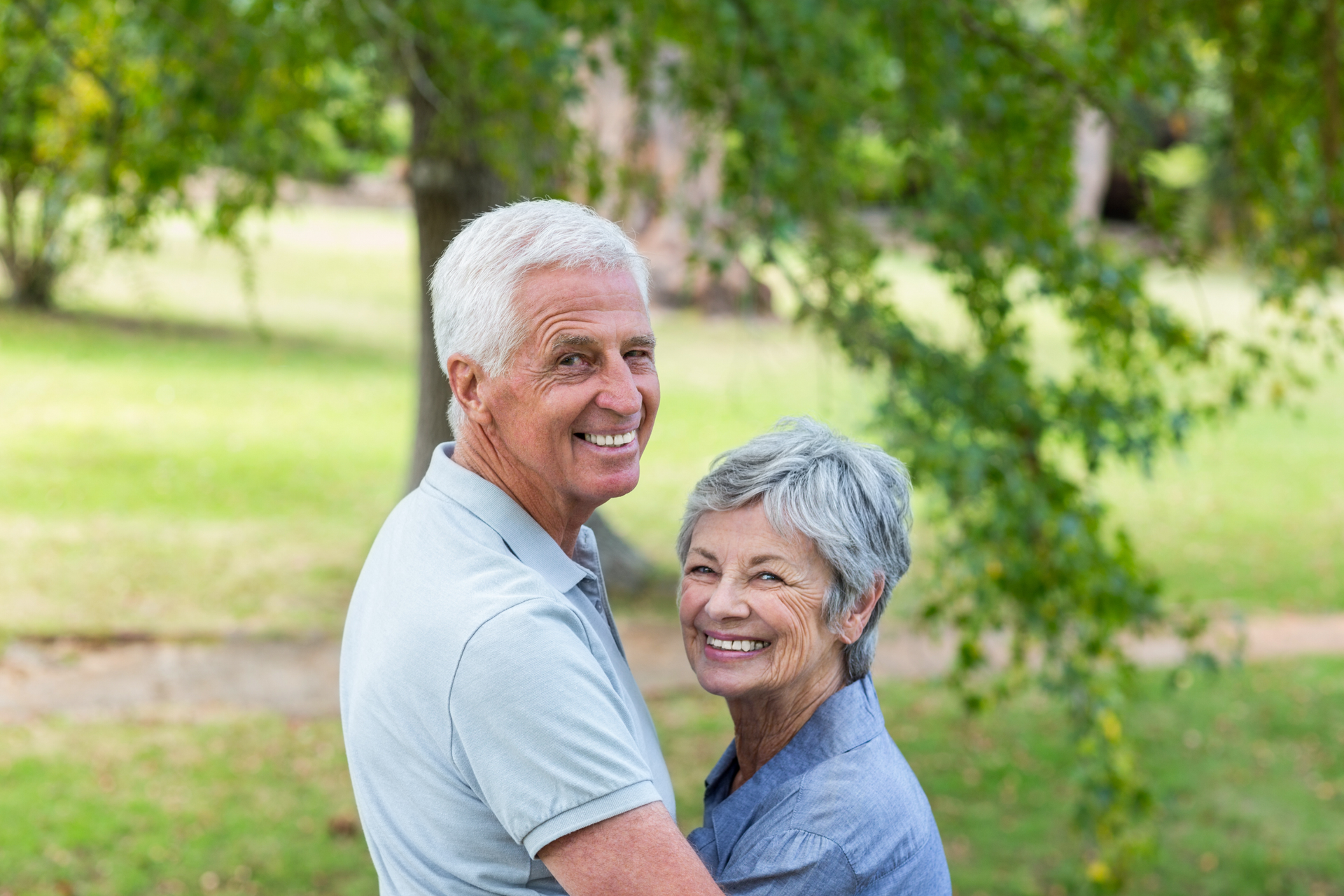 Smiling elderly couple embracing outdoors in a park, surrounded by trees and greenery.