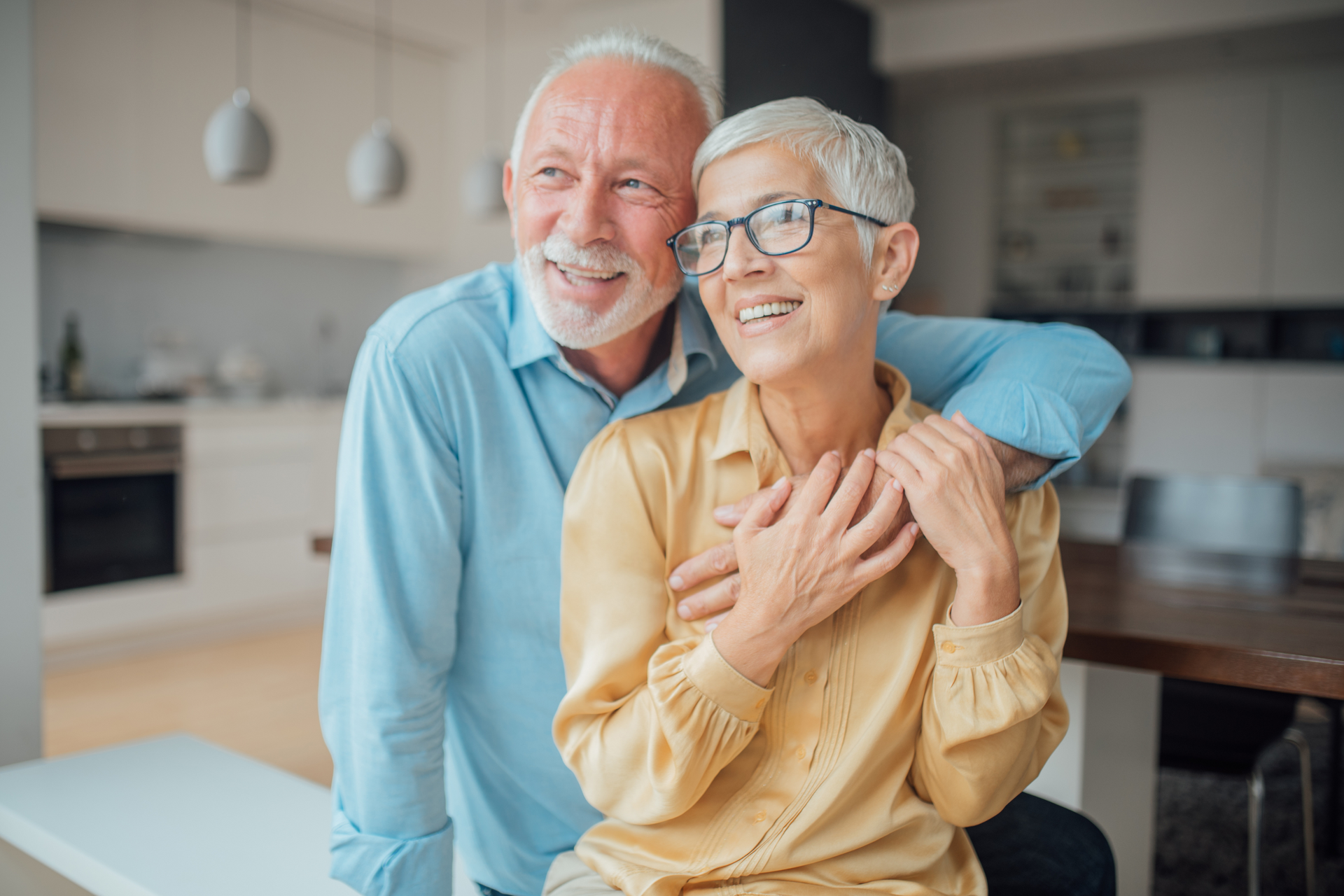 Senior couple embraces indoors, man's arm around the woman's shoulders. They smile, looking out of frame in a bright kitchen.