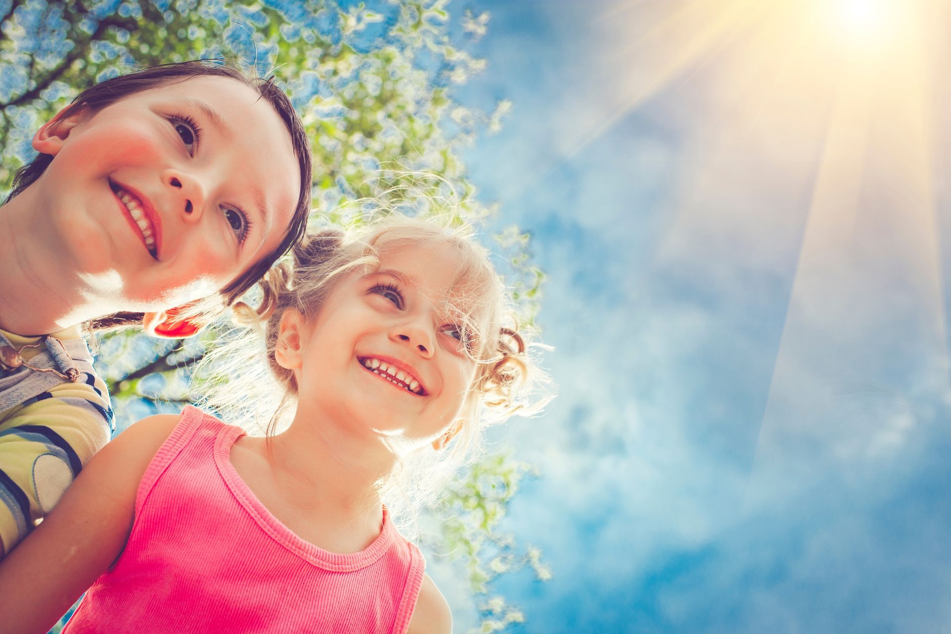 Two smiling children looking up at the bright sun in a blue sky, with trees visible in the background.