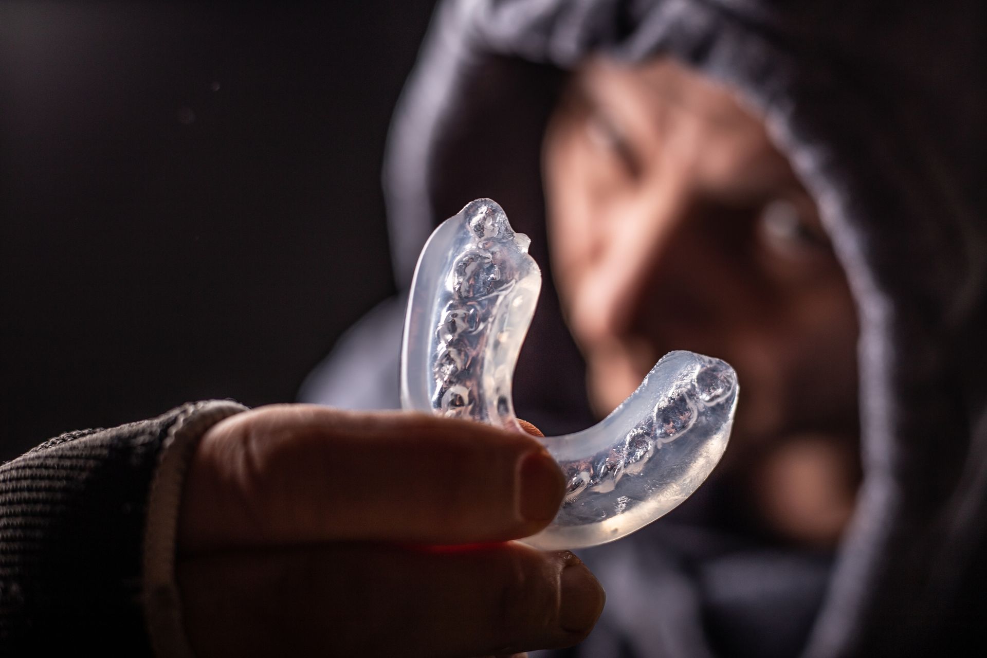 Person holding a clear, translucent mouth guard, looking towards it. They are in a hoodie against a dark background.