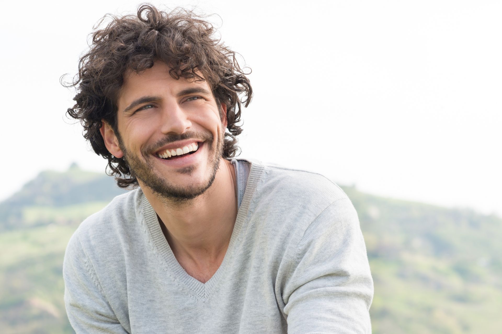 Man with curly hair smiles broadly outdoors, wearing a gray sweater, with a blurred green background.