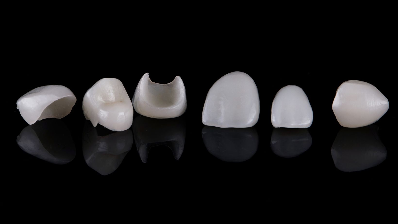 Six porcelain dental veneers, white and curved, arranged against a reflective black background.