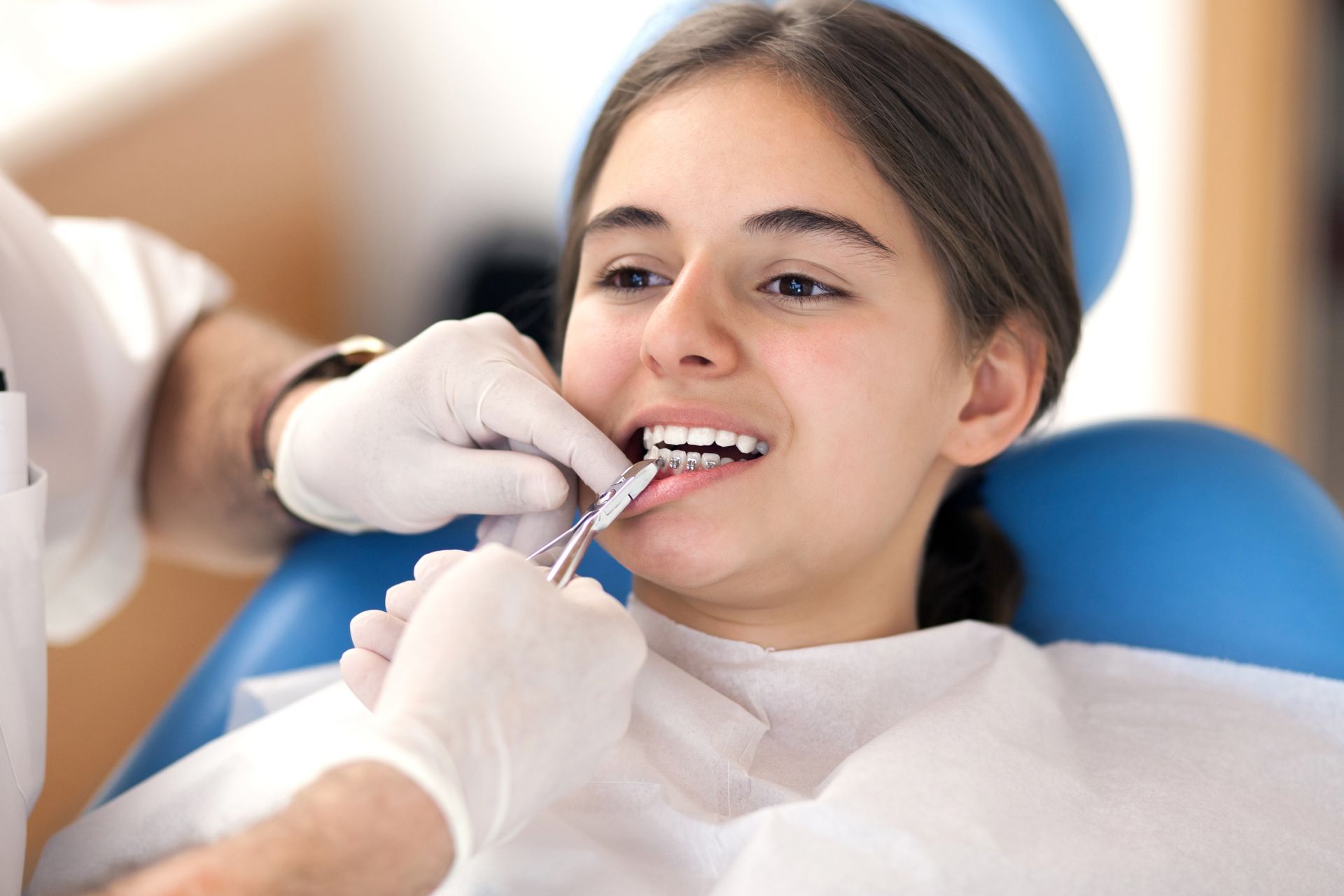 A dentist examines a young woman's teeth with a tool in a dental chair. Both are wearing gloves.