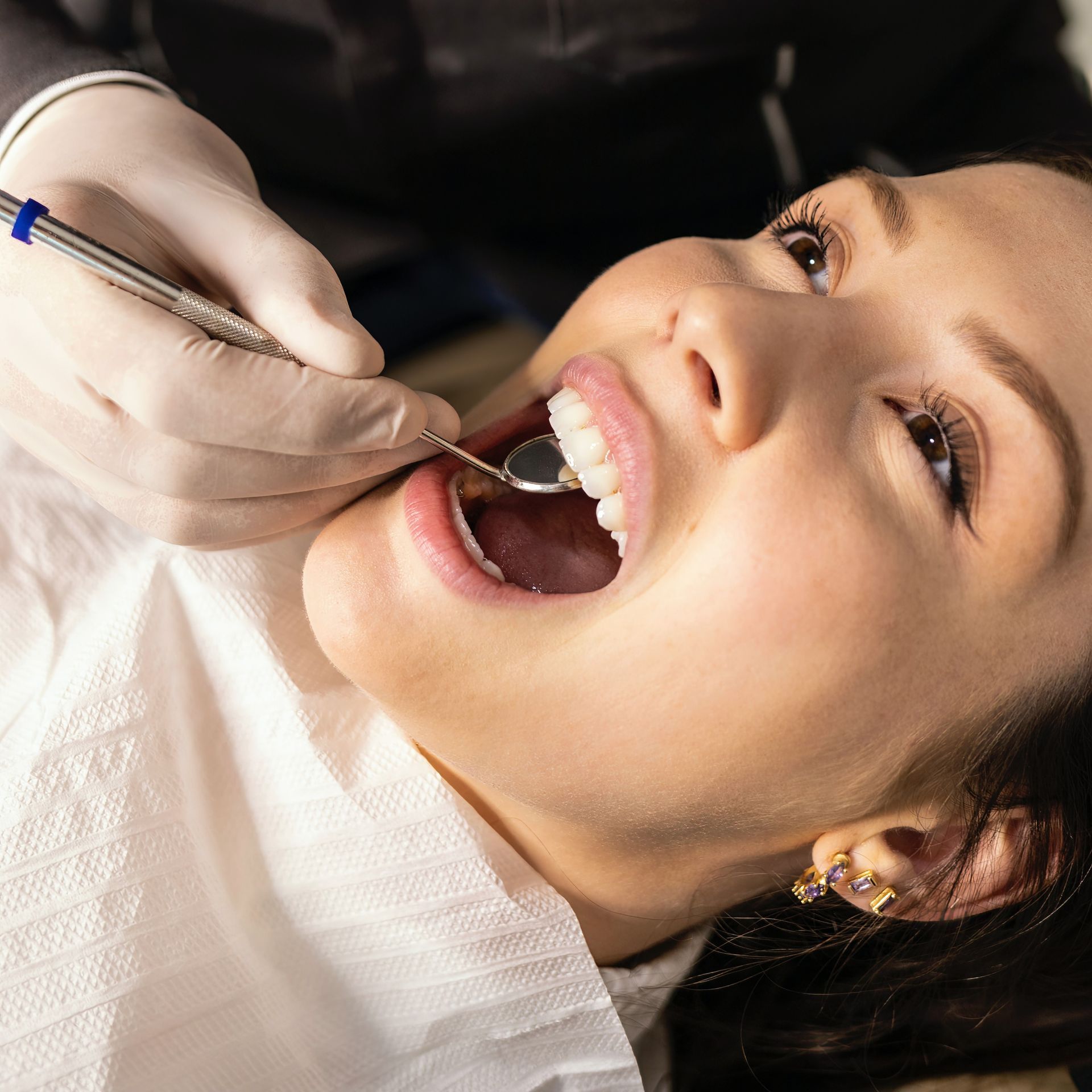 A dentist examining a woman's teeth. The woman lies back with her mouth open, a dental mirror reflecting her teeth.