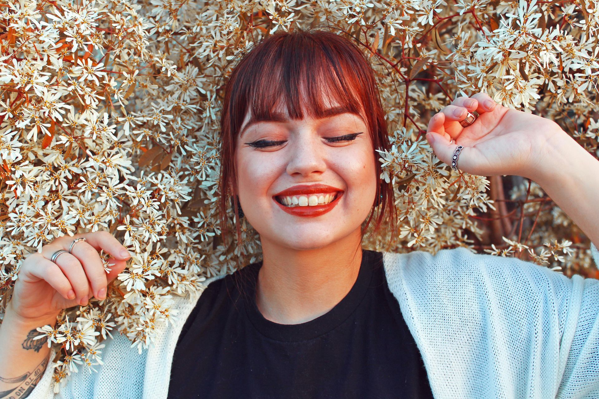 Woman with red-tinted hair and dark lipstick smiles brightly, leaning against a flowering bush with white blossoms.