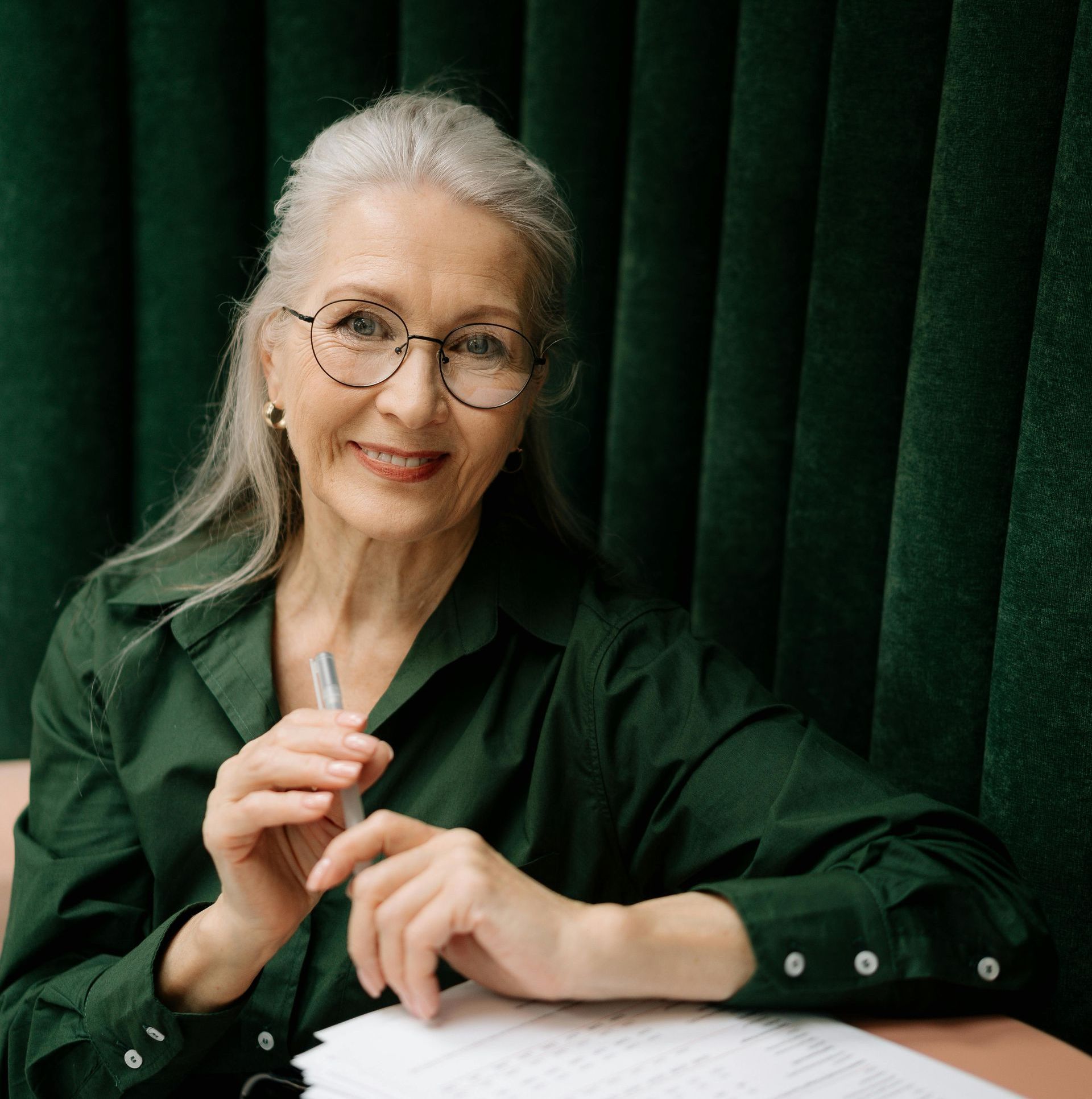 Smiling older woman with gray hair, wearing glasses and a green shirt, holding a pen, seated at a table with papers.