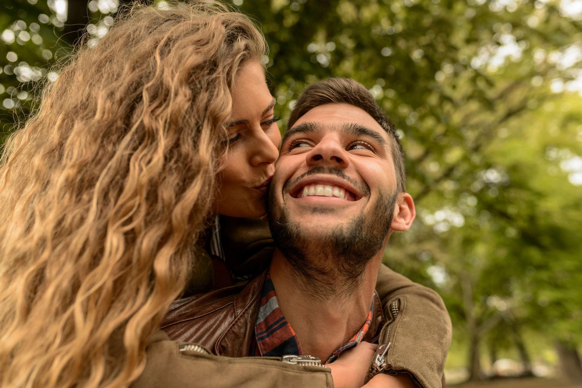 Woman kisses a smiling man on the cheek outdoors; they are embracing with a blurred green tree backdrop.
