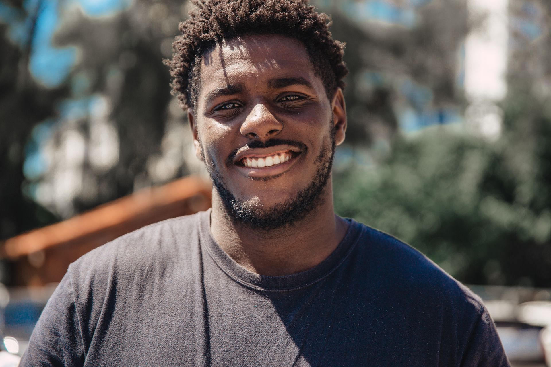 Smiling Black man with short curly hair, wearing a black t-shirt, in an outdoor setting with trees in the background.