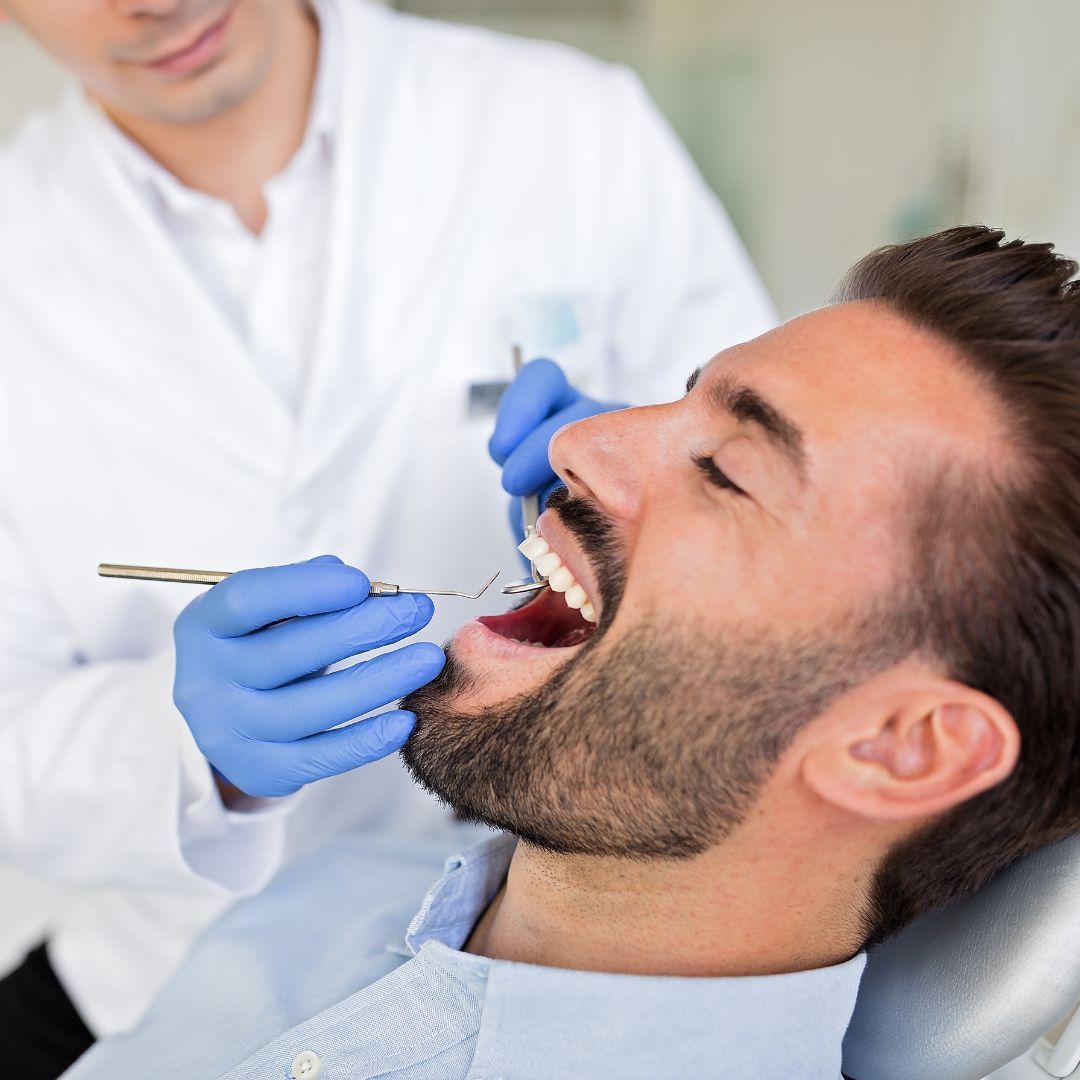 A dentist examines a man's teeth in a clinic. The man sits in a chair with his mouth open.