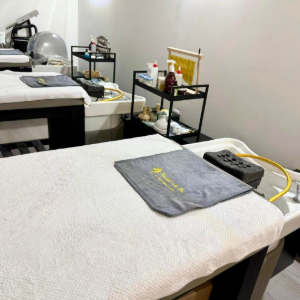Massage room with two treatment tables, gray towels, and small black carts against white walls.