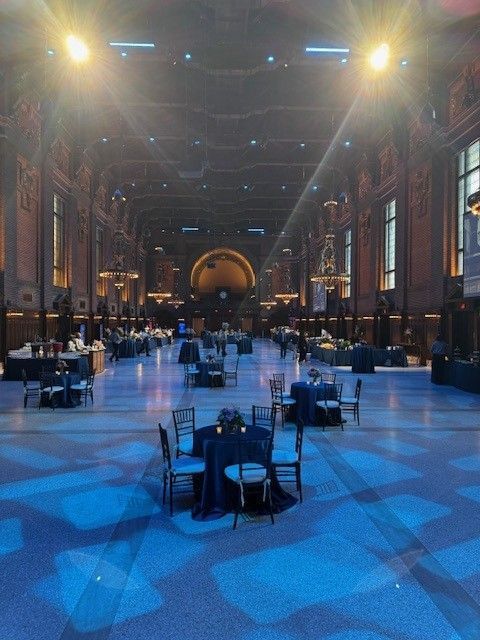 Large event space with tables, chairs, and blue lighting. Ornate architectural details and a high ceiling are visible.