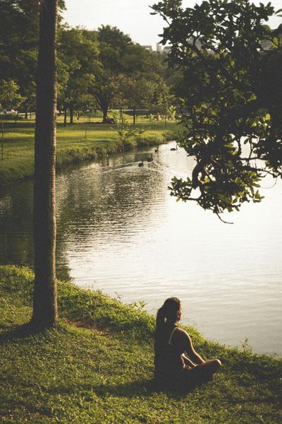 Woman meditating by a calm pond in a park, surrounded by green grass and trees. Sunlight filters through.