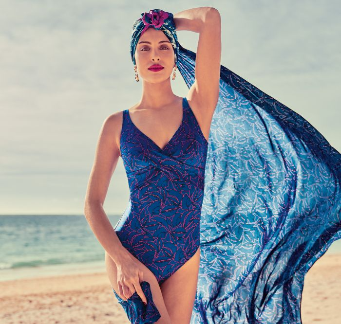 A woman in a blue swimsuit is holding a blue scarf on the beach.