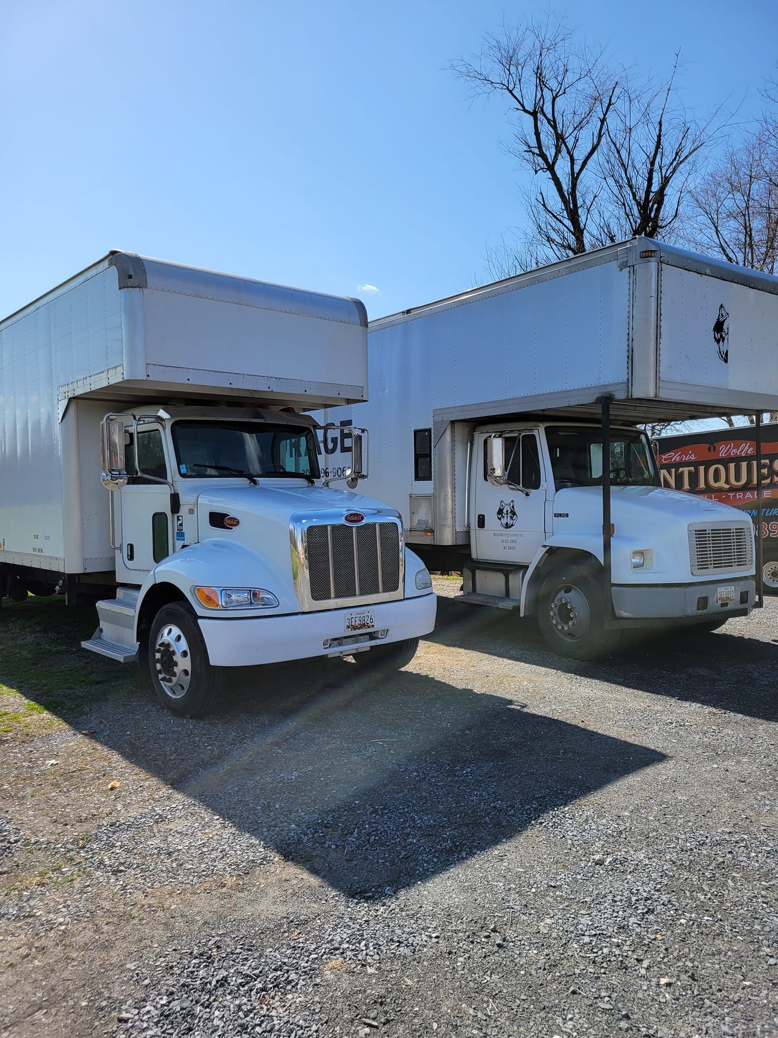 Two white moving trucks are parked next to each other in a gravel lot.