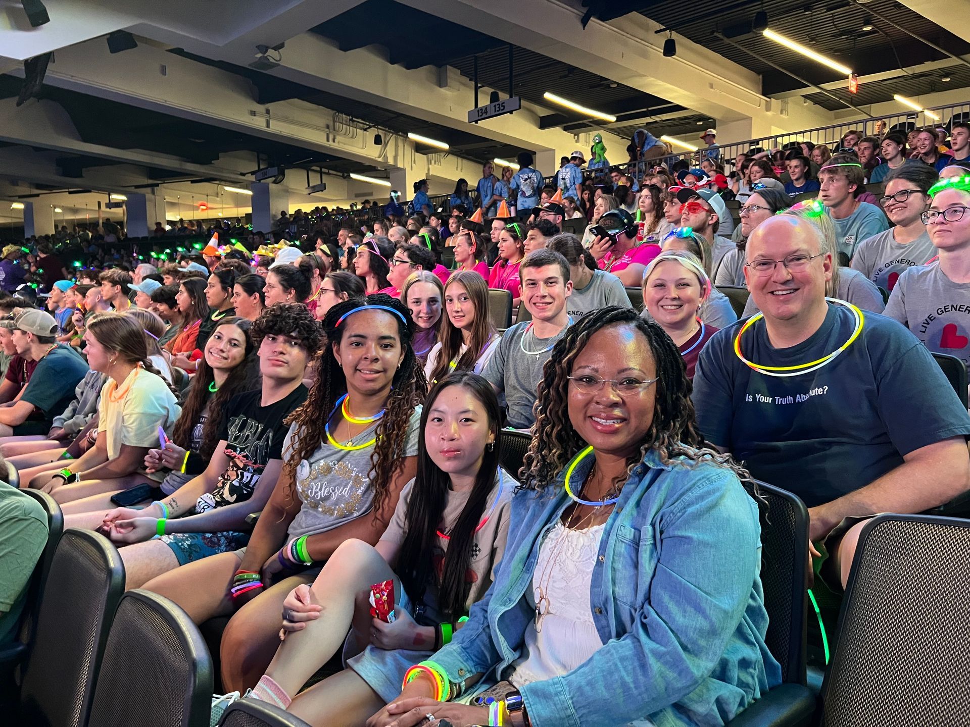 People in a crowded auditorium, wearing glow sticks. Smiling faces; several people in the foreground.