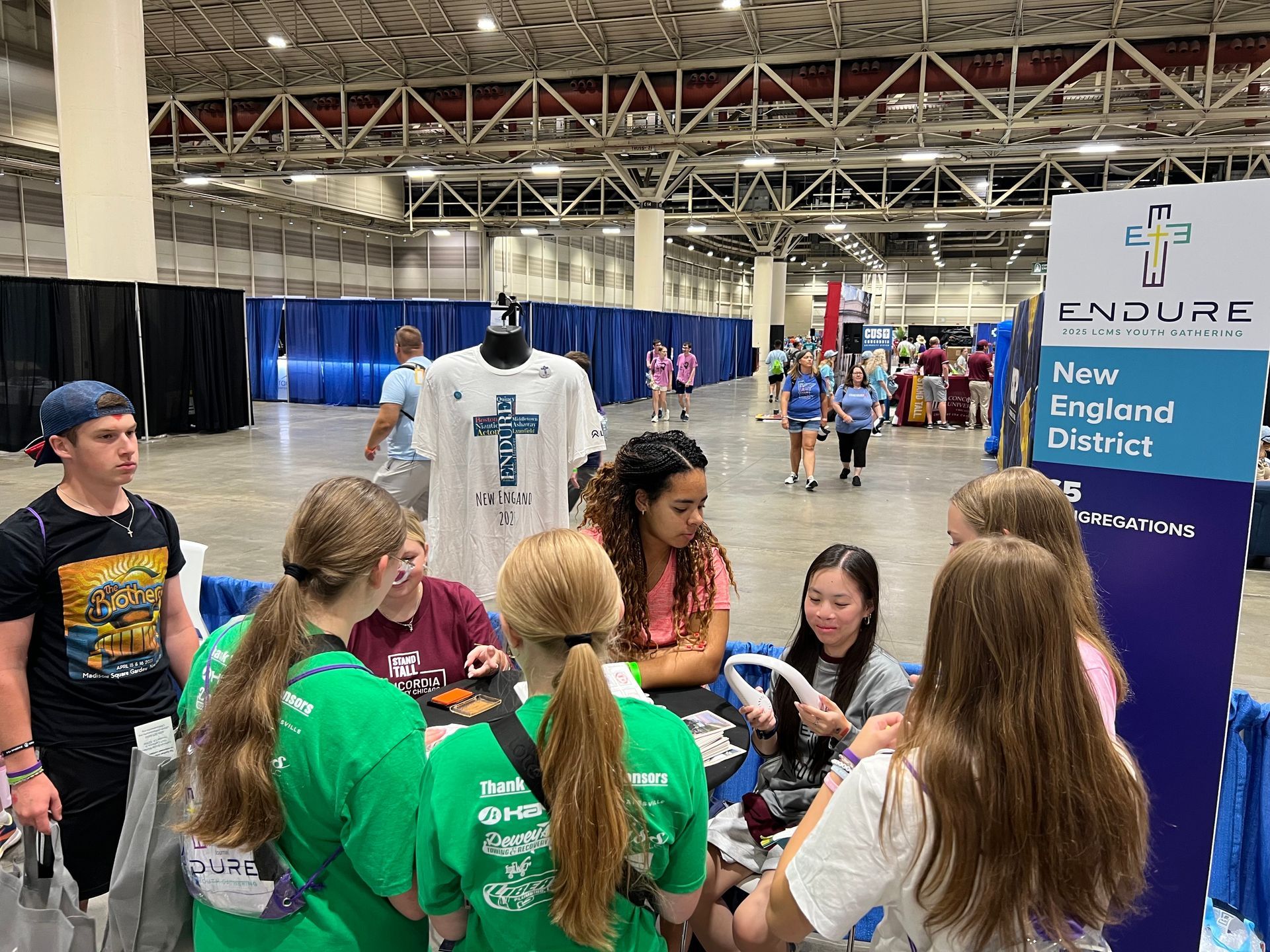 People at a booth for the New England District. A shirt with a cross is on display.