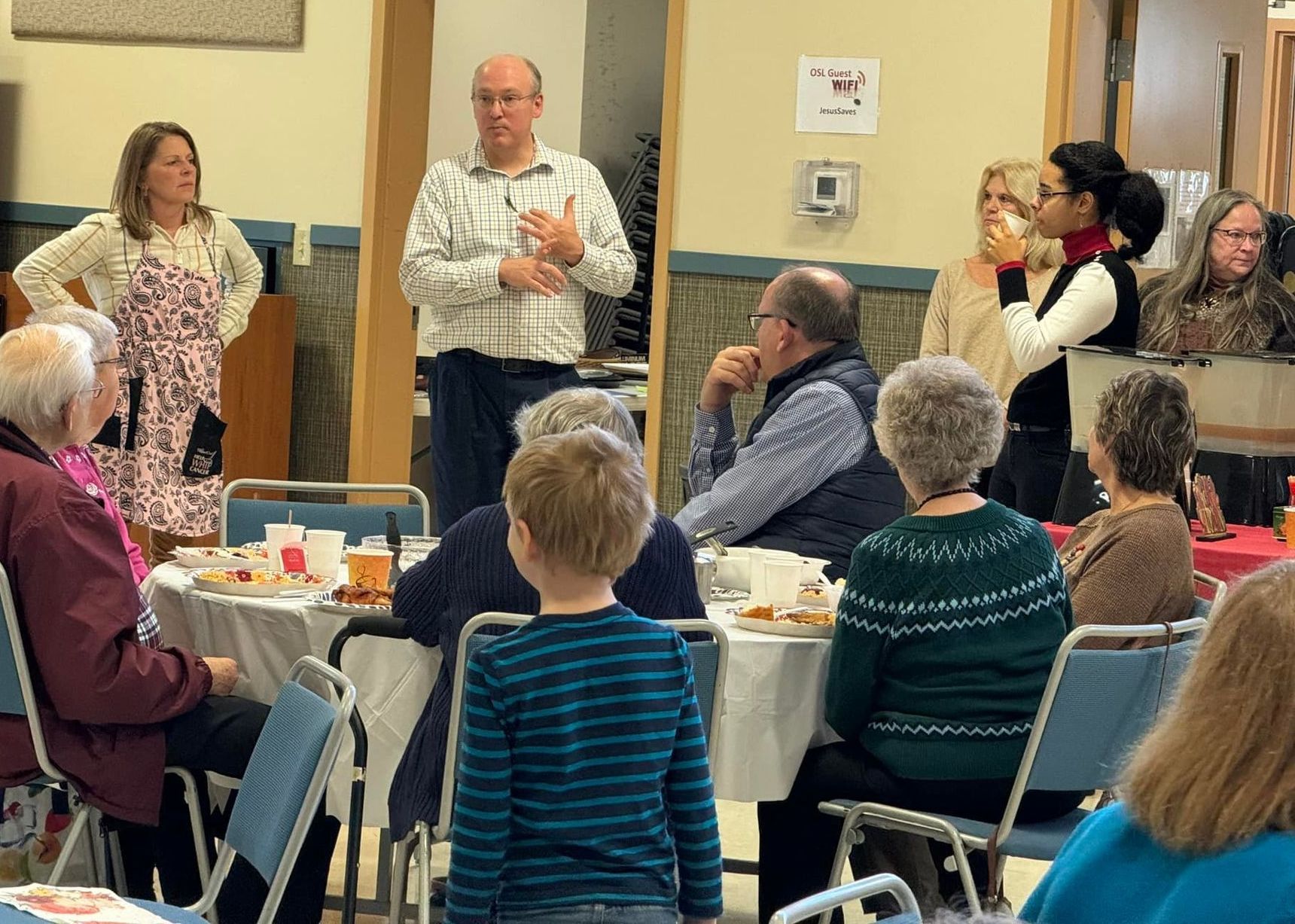 People gathered for an event. A man speaks while others listen. Tables are set with food and drinks.