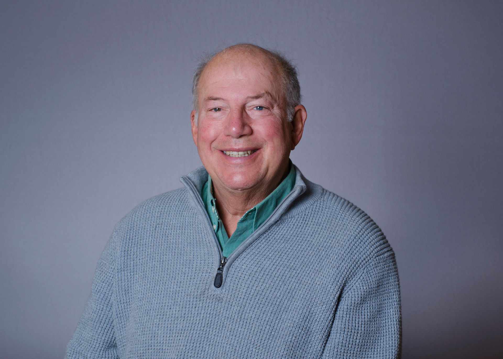 Smiling older man in blue sweater against a grey background.