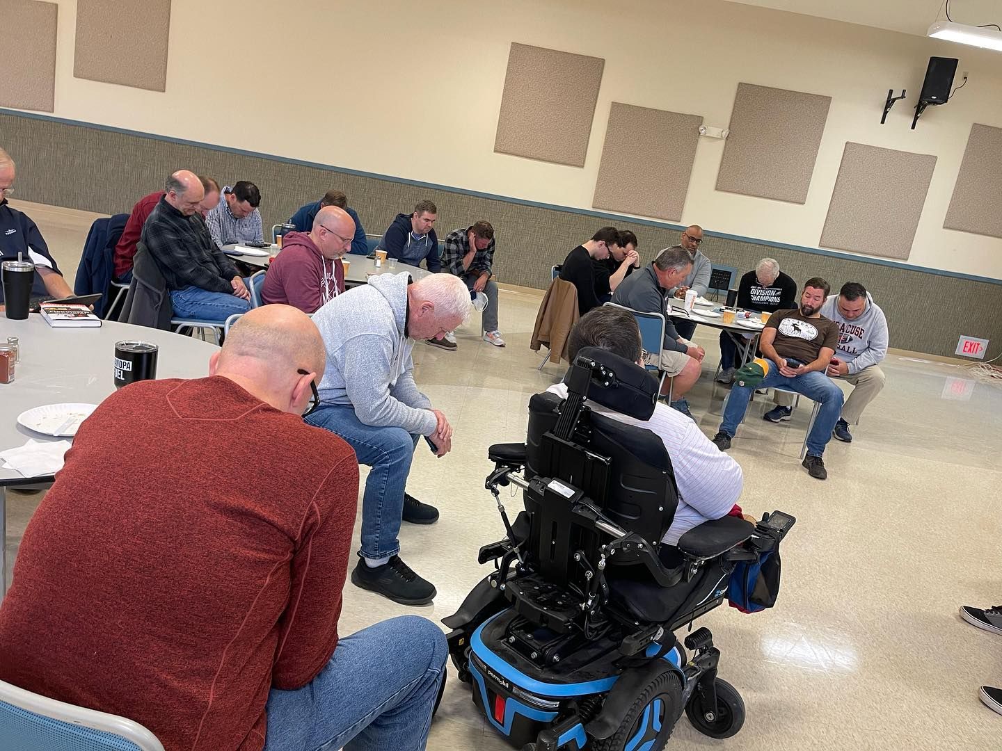 Men in a meeting, some seated at tables, one in a wheelchair. Beige room with soundproofing panels.