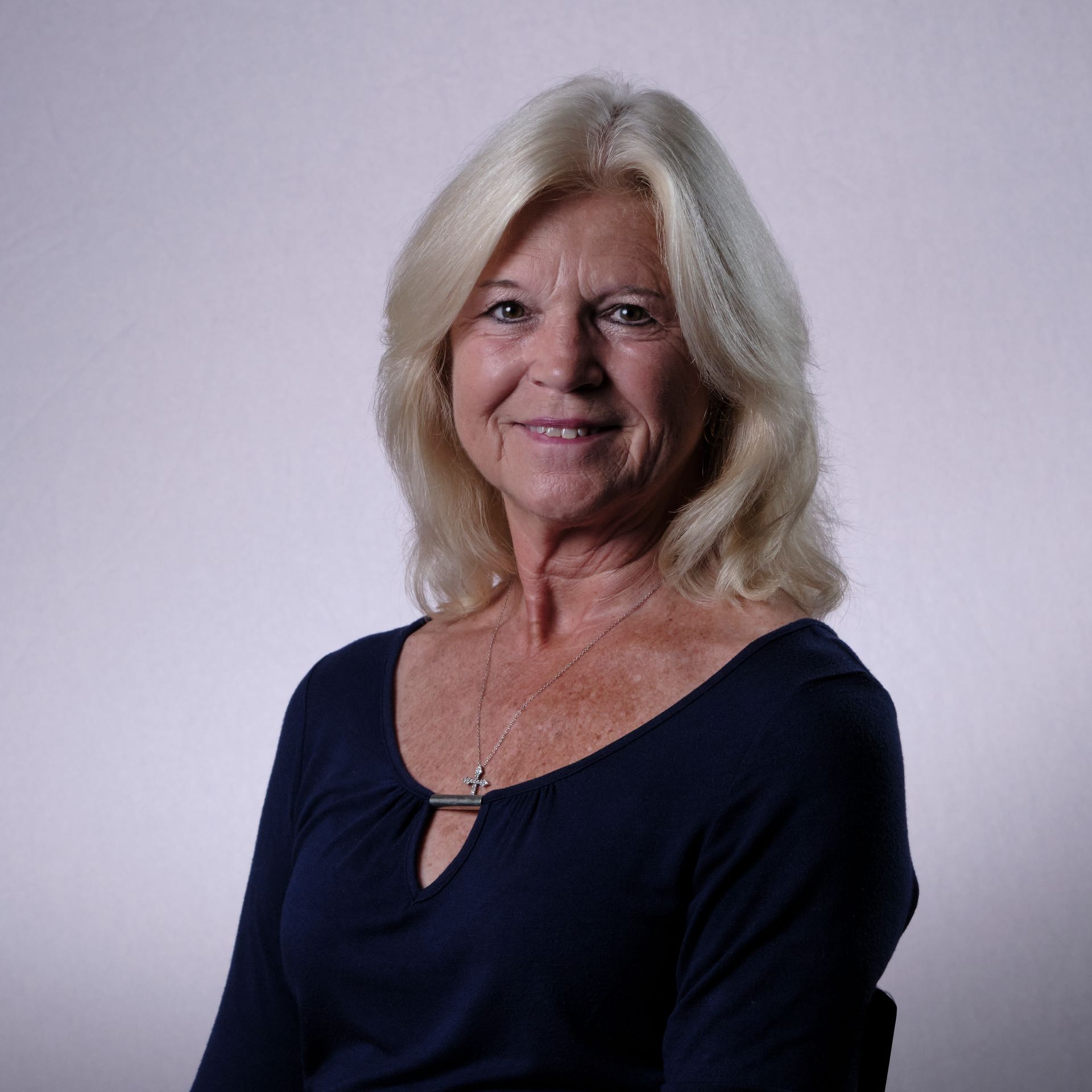 Blonde woman with a warm smile in a dark blue top, looking at the camera, against a light background.