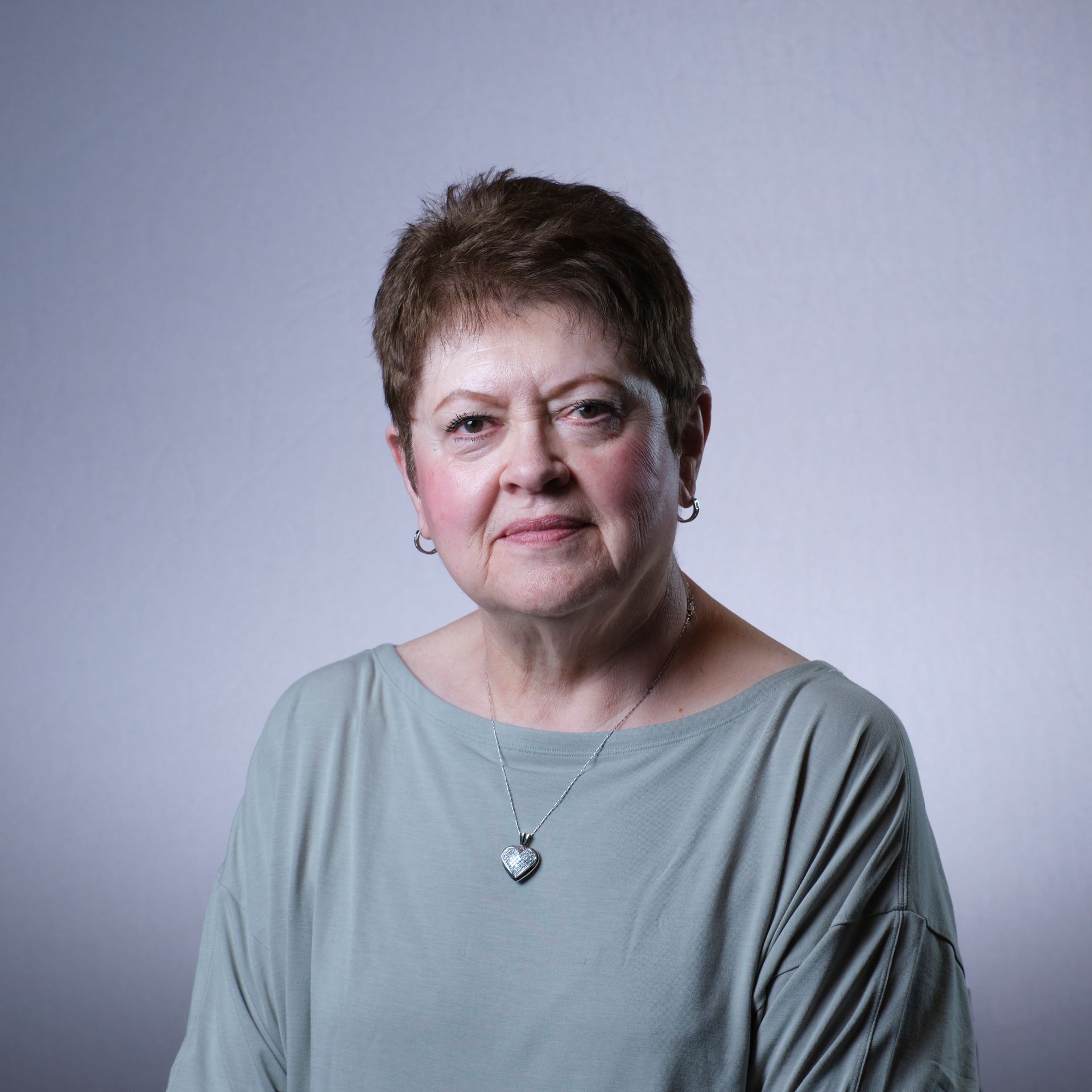 Woman with short brown hair wearing a green top and a silver heart pendant against a light gray background.