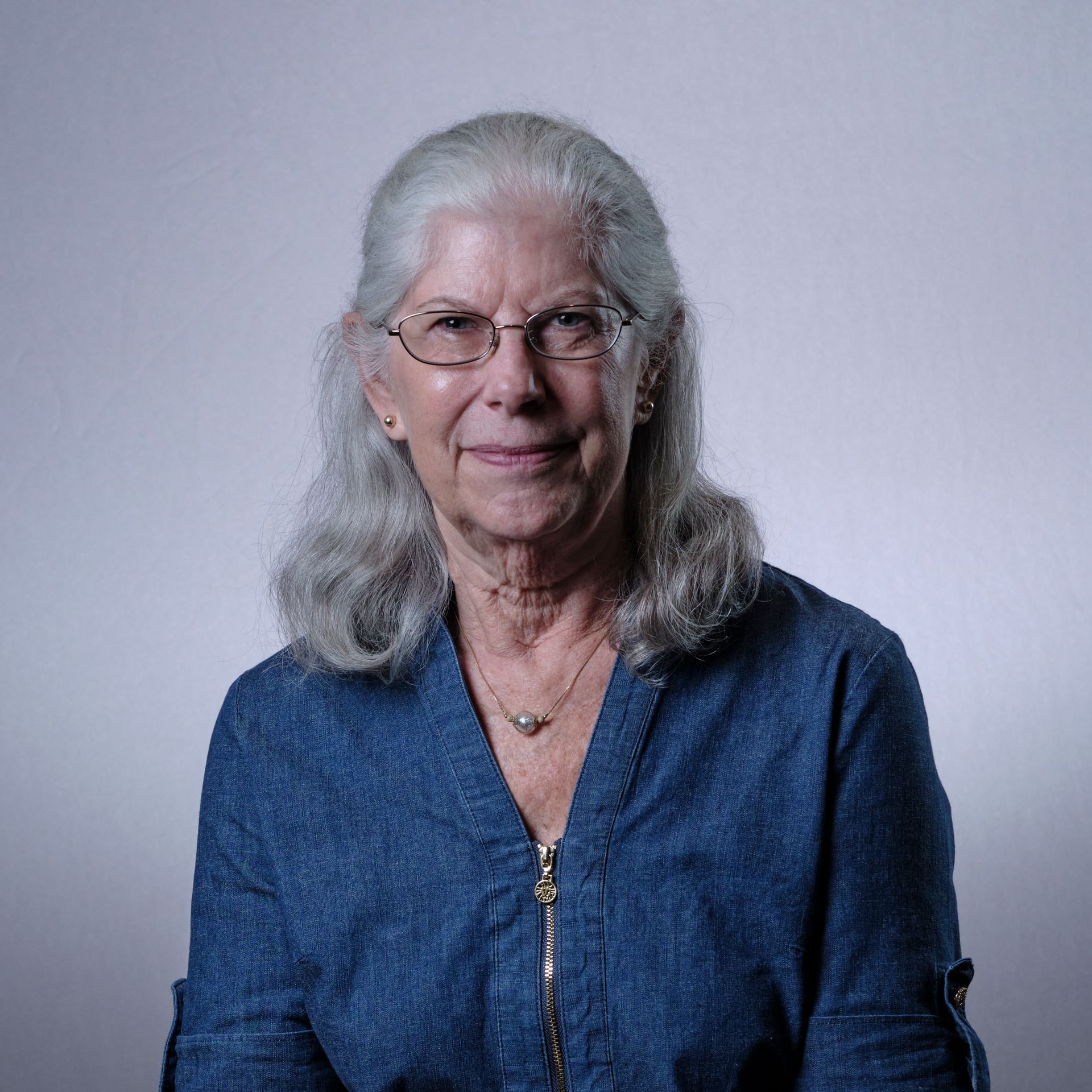 Woman with gray hair, glasses, and blue shirt, smiles at the camera.