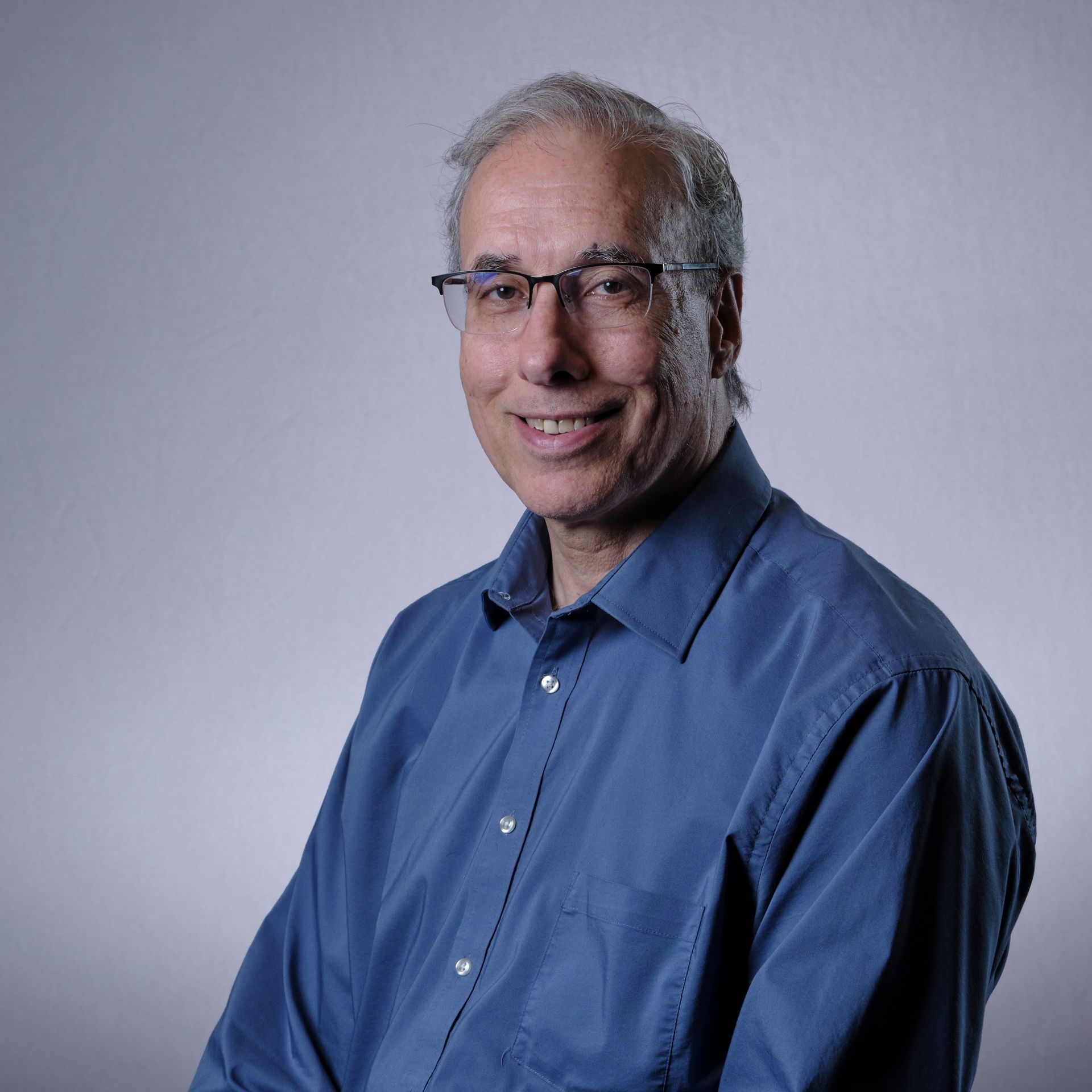 Man with glasses smiles at the camera, wearing a blue shirt, against a gray backdrop.