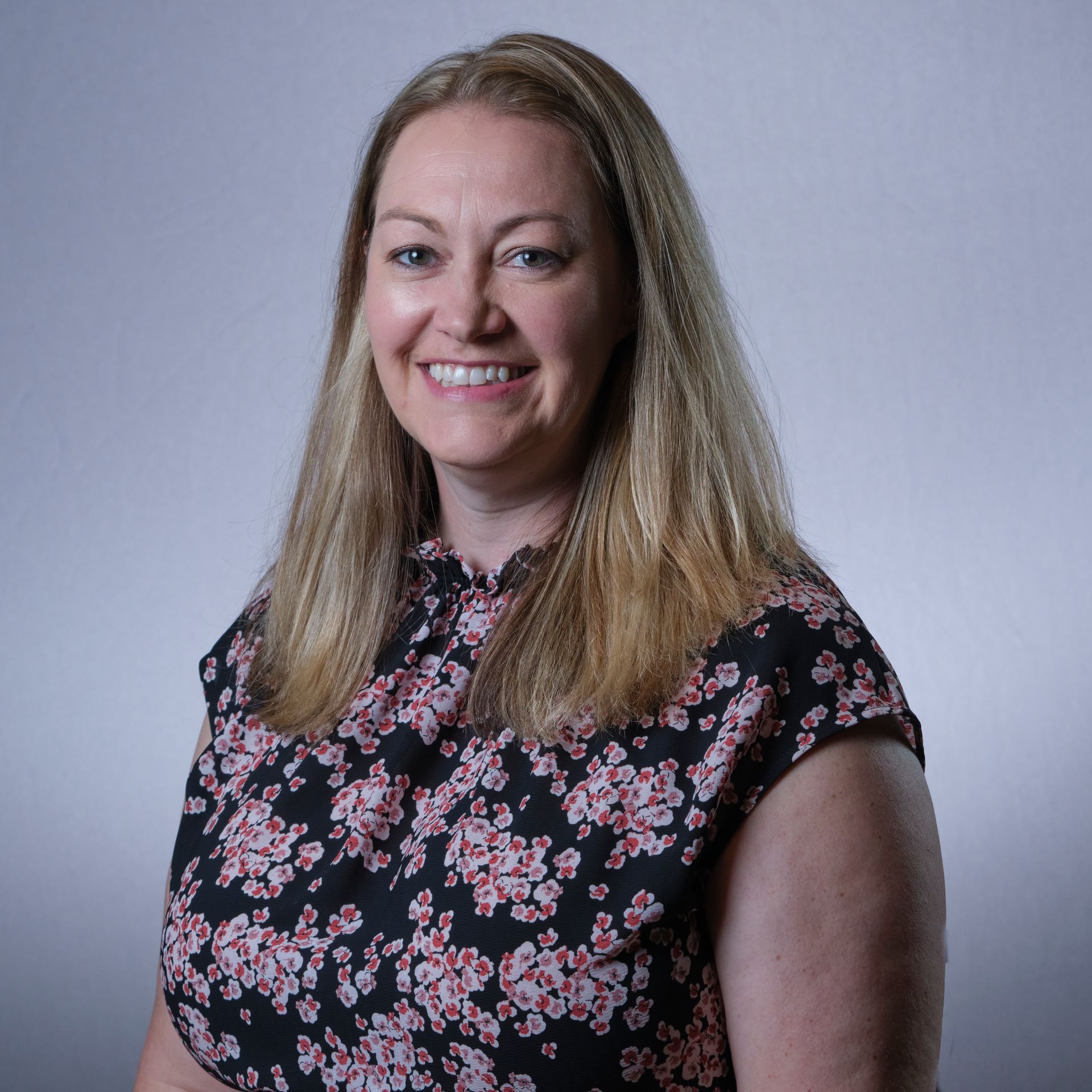 Woman with blonde hair, smiling, wearing a floral top, against a neutral background.