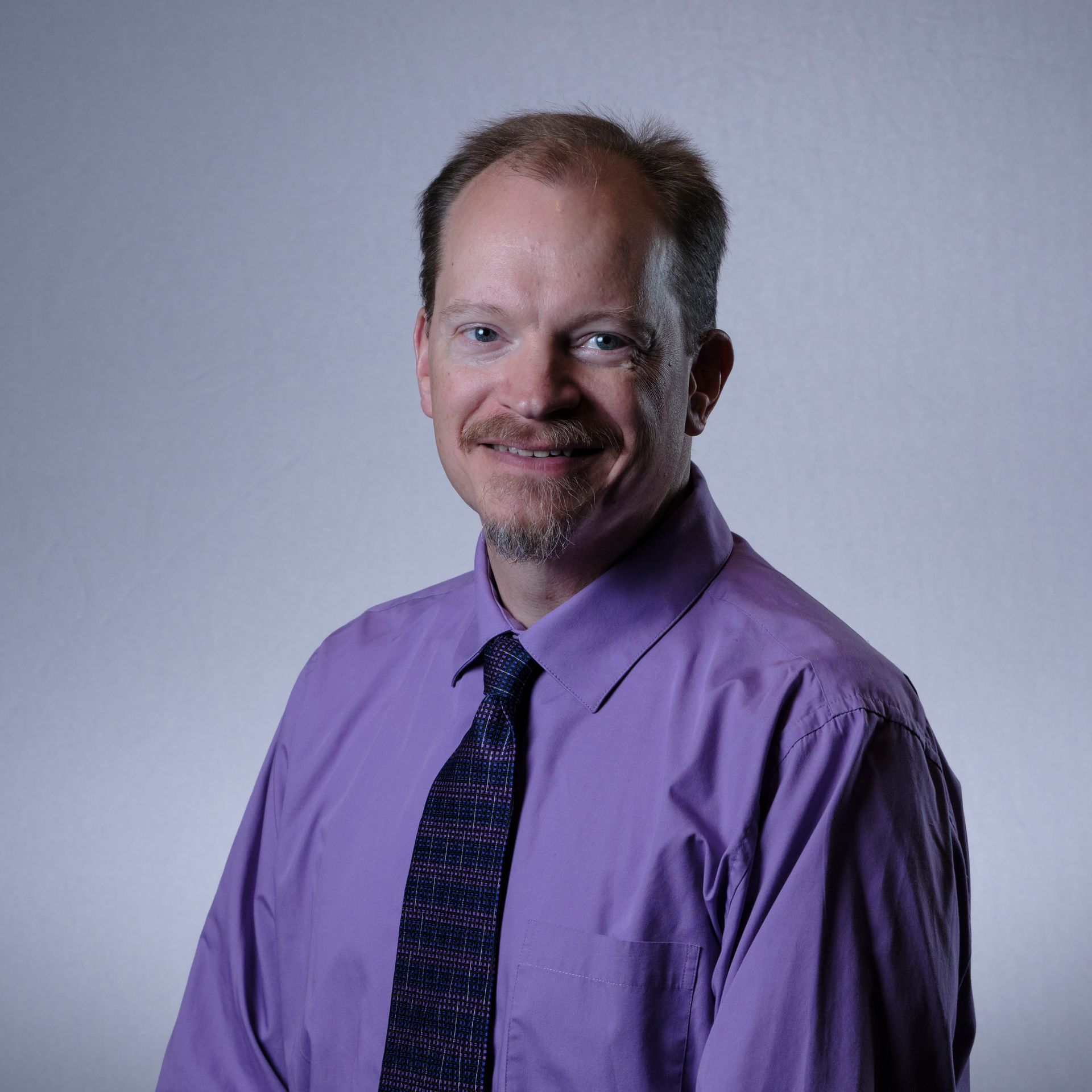 Man with fair skin, wearing a purple shirt and tie, smiling against a grey background.