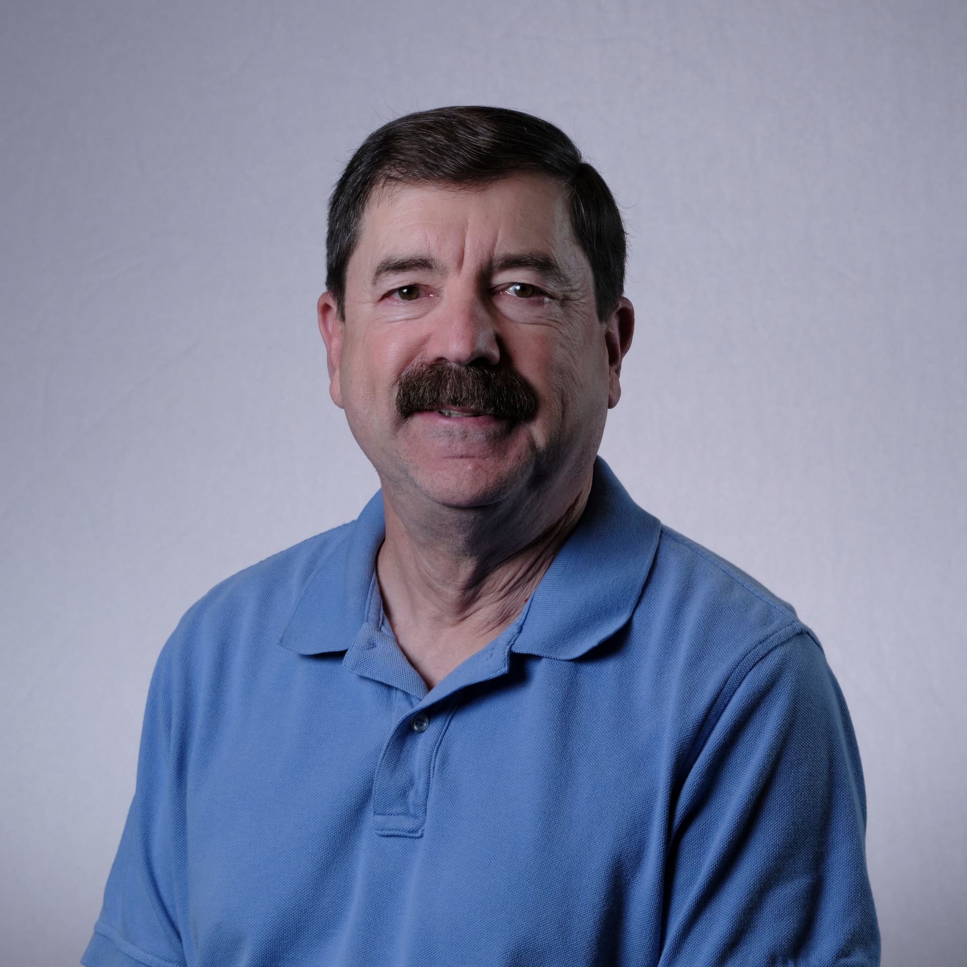 Man with dark hair and mustache in a blue polo shirt, smiling. White background.