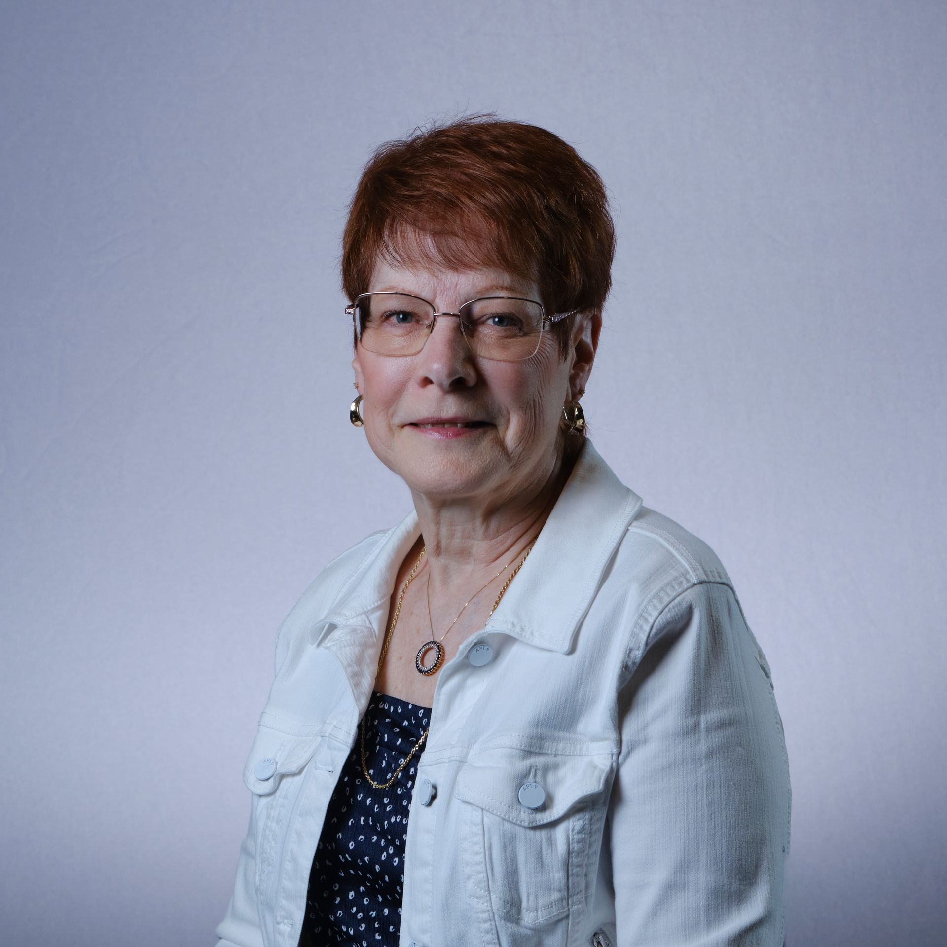 Woman with short red hair, glasses, white jacket, and polka dot top smiles.