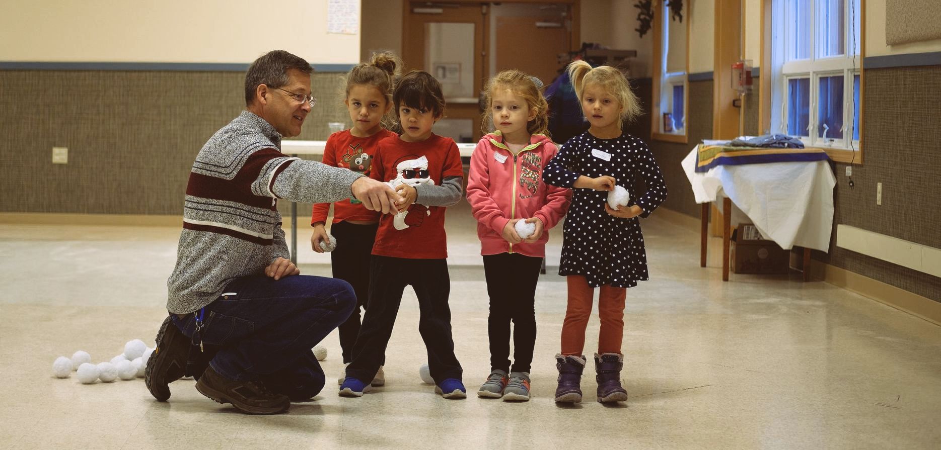 A man demonstrates something to four children in a room. The children hold balls.