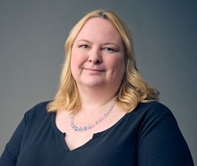 A person with shoulder-length blonde hair wearing a dark blue v-neck shirt and a beaded necklace against a gray background.