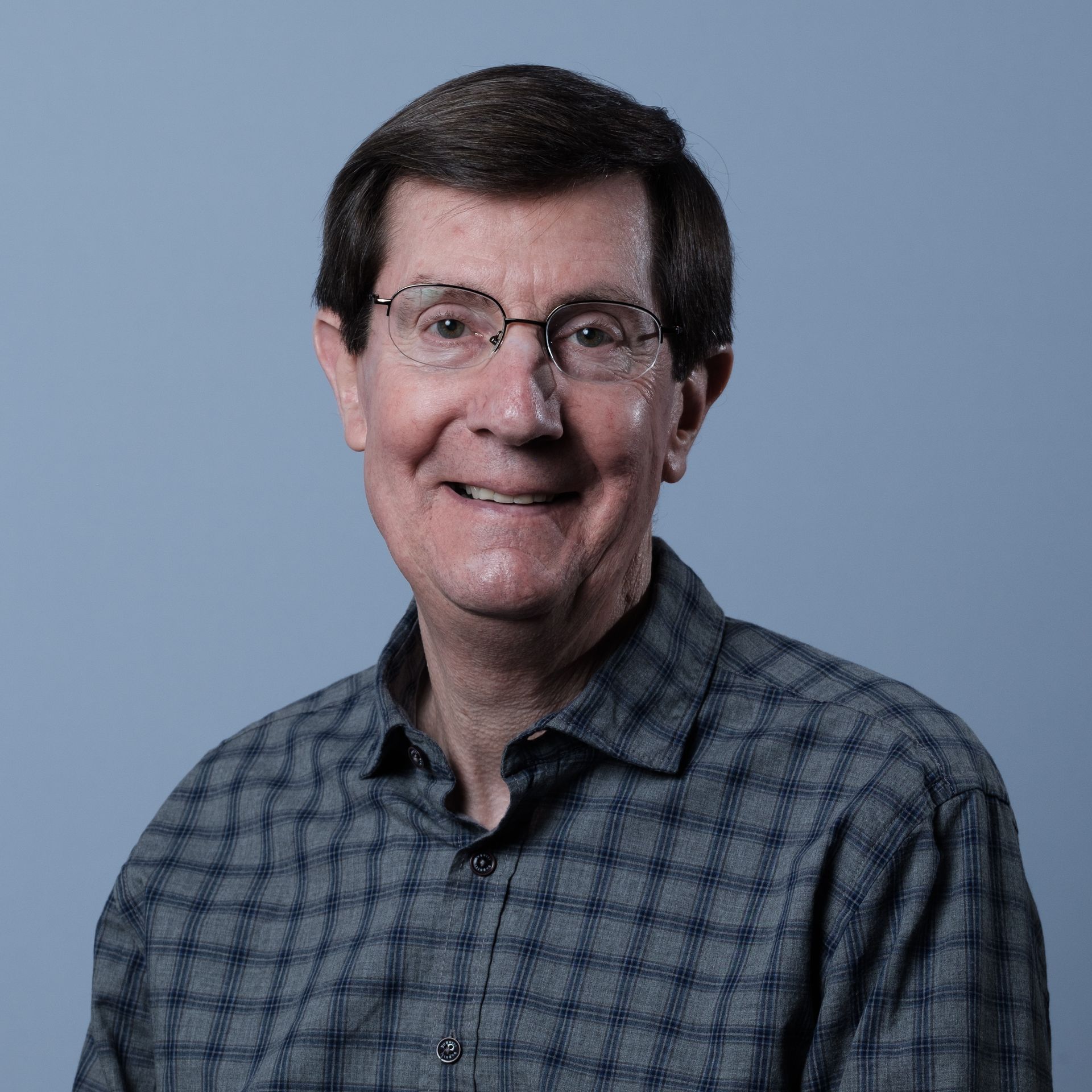 A man with glasses smiles, wearing a blue plaid shirt, set against a blue background.