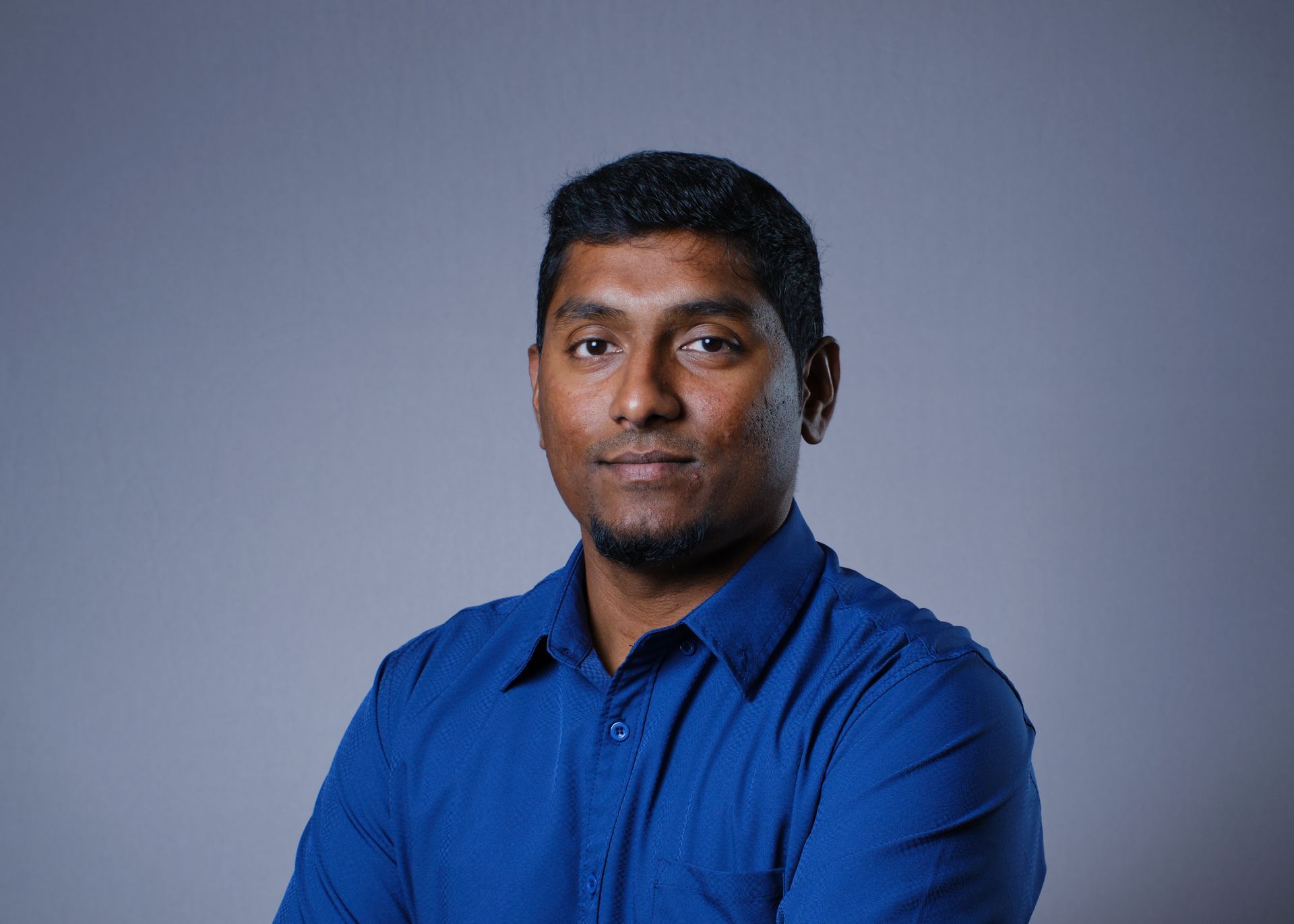 Man with dark hair and beard, wearing a blue shirt, arms crossed, looking at the camera.