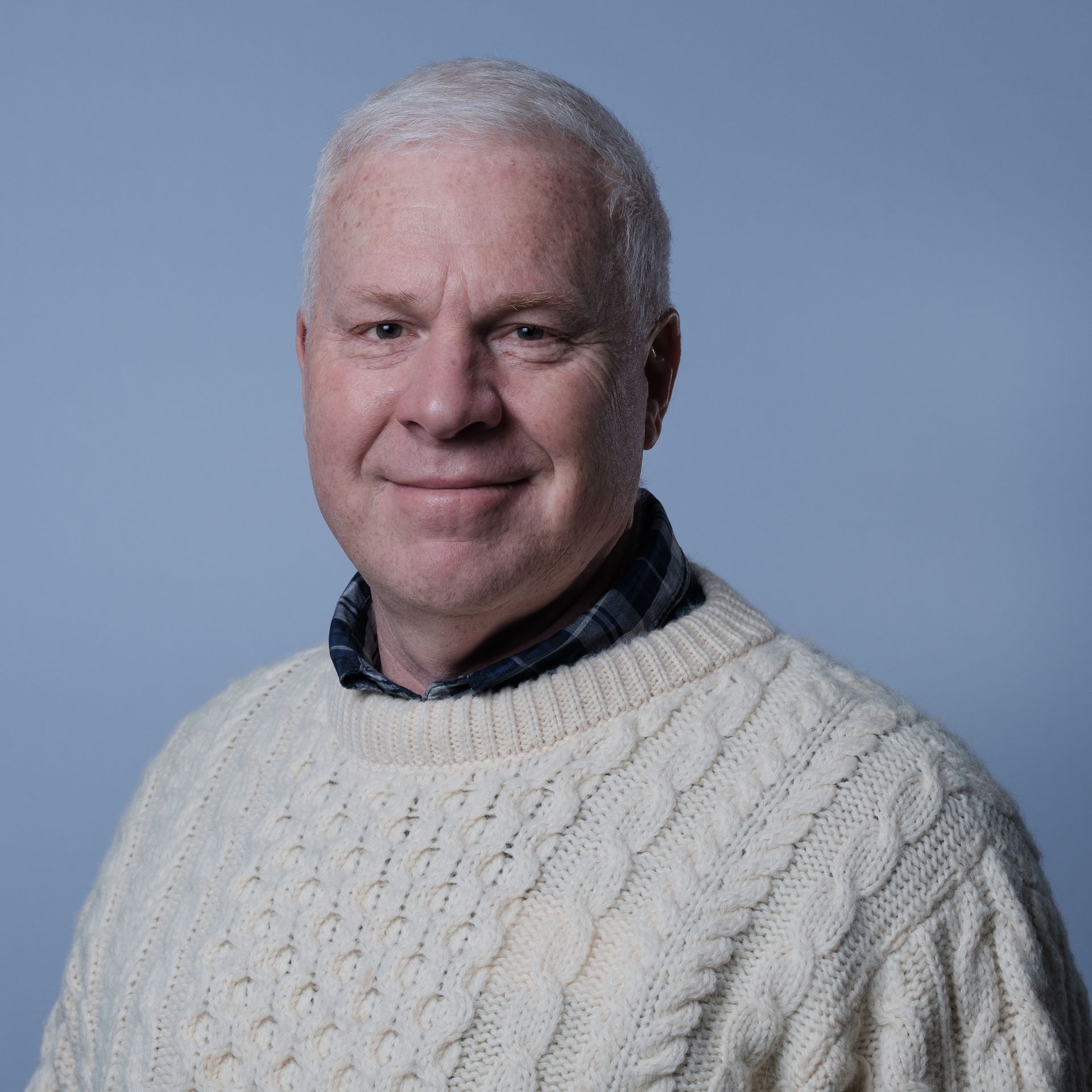Man with gray hair, wearing a white cable-knit sweater and plaid shirt, smiles, facing the camera.