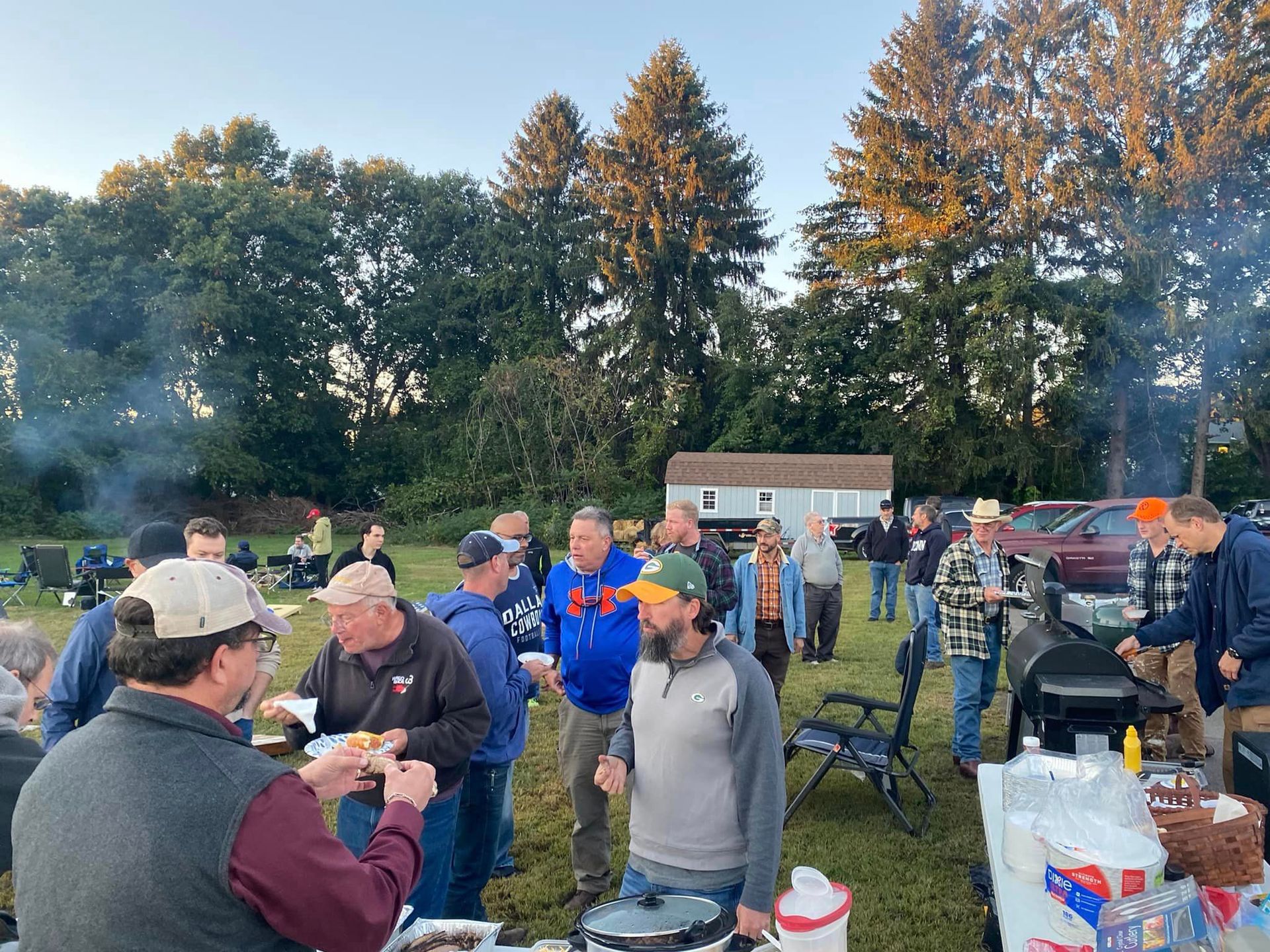 Outdoor gathering: people at a barbecue. Men in casual clothes are gathered, some eating, with trees and a small building in the background.