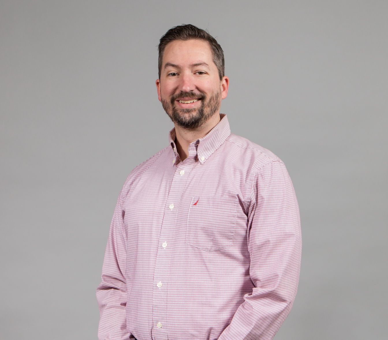 Man with a beard wearing a pink and white striped button-down shirt, smiling at the camera. Grey background.