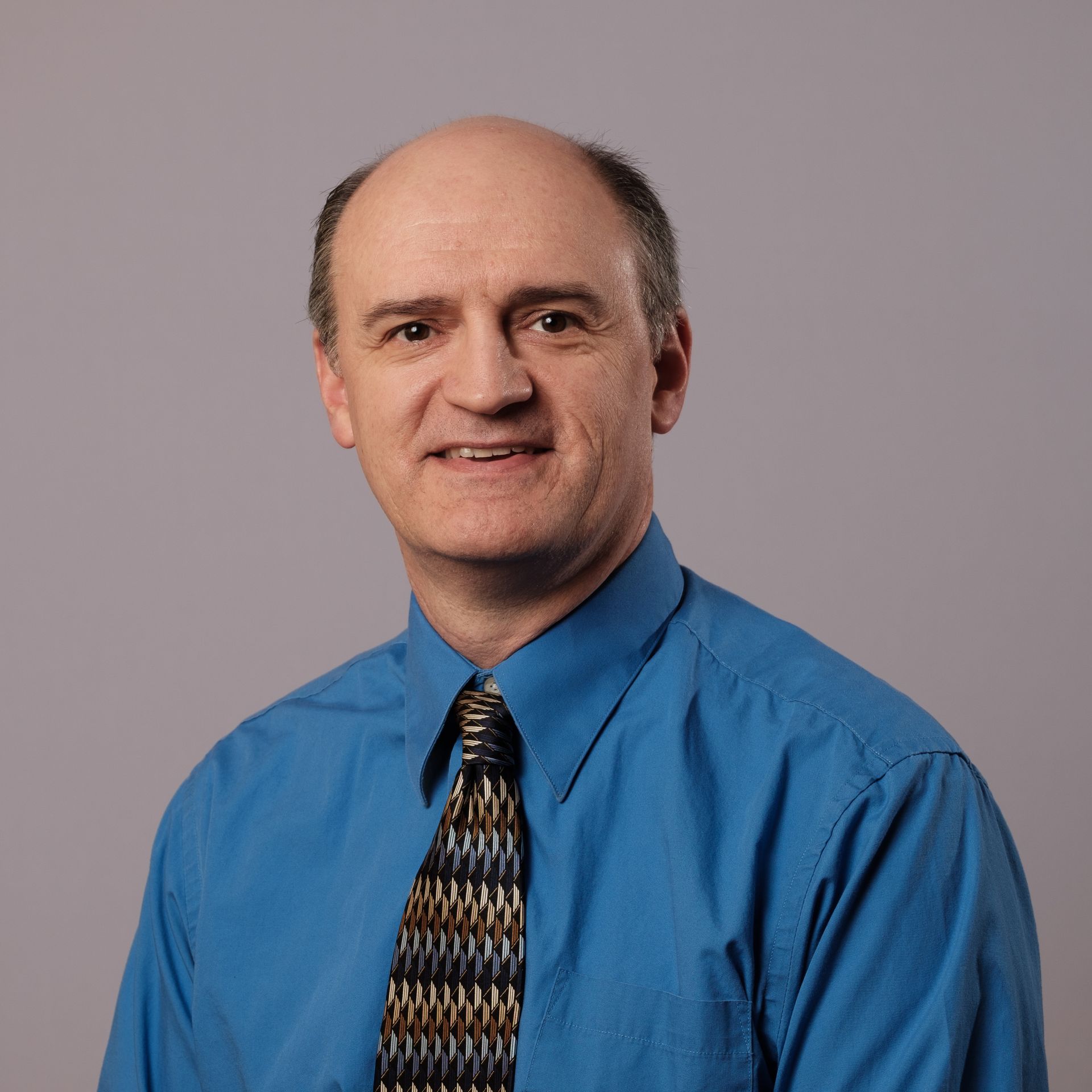 Bald man in blue shirt and patterned tie smiles at the camera, against a gray background.