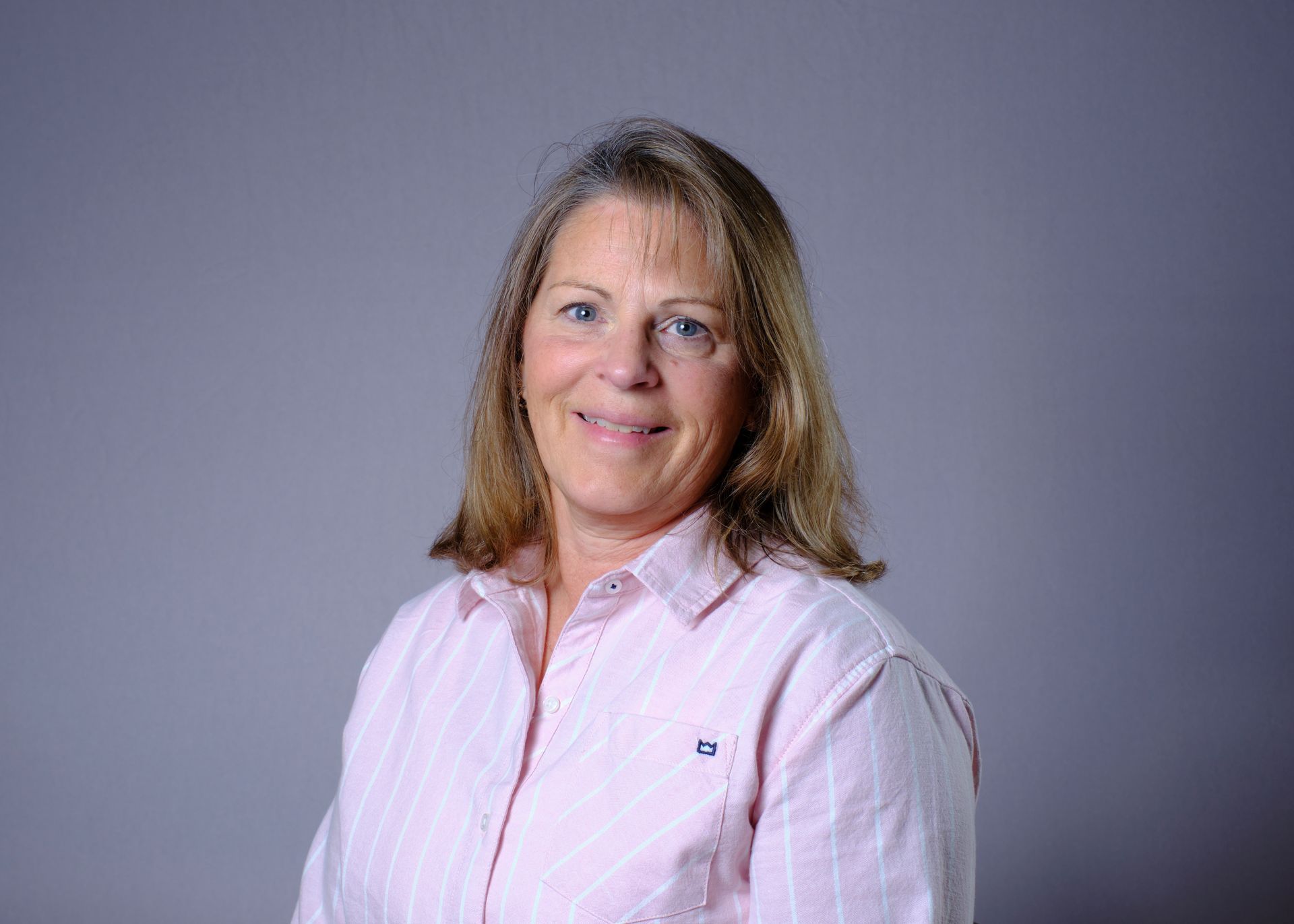 Woman with fair skin smiles, wearing a pink striped button-down shirt, standing against a gray background.