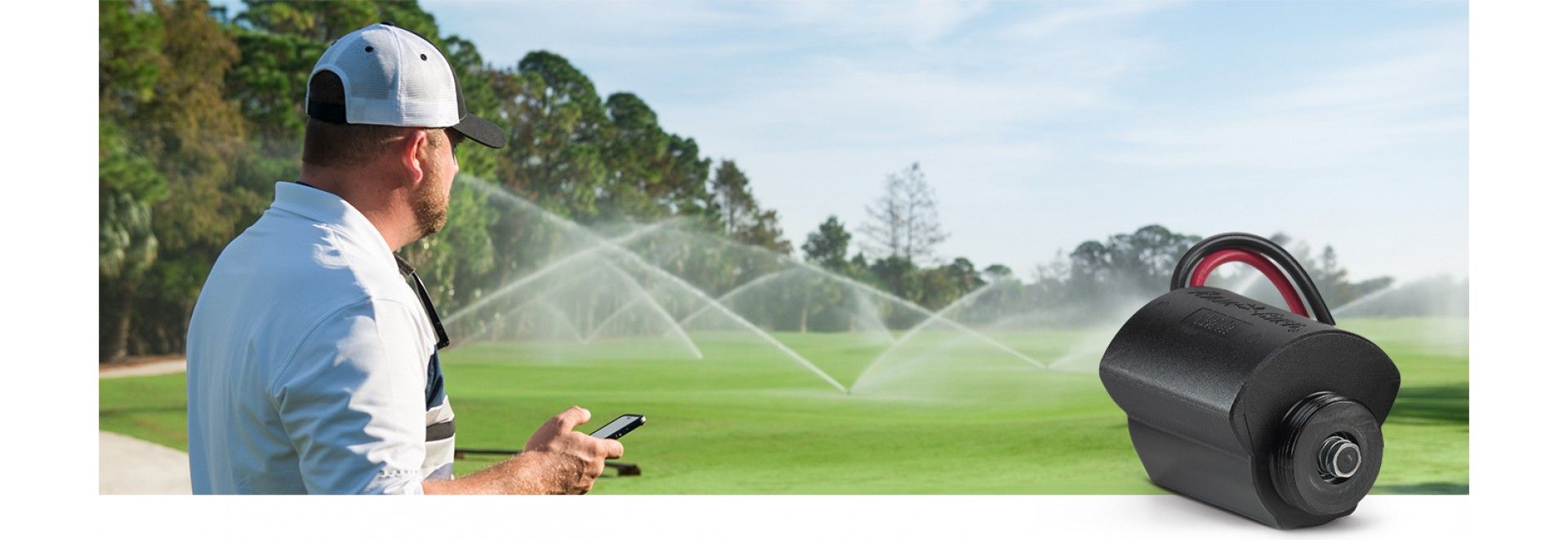 A man in a white hat observes a golf course being watered with a sprinkler system. A black cylinder is in the foreground.