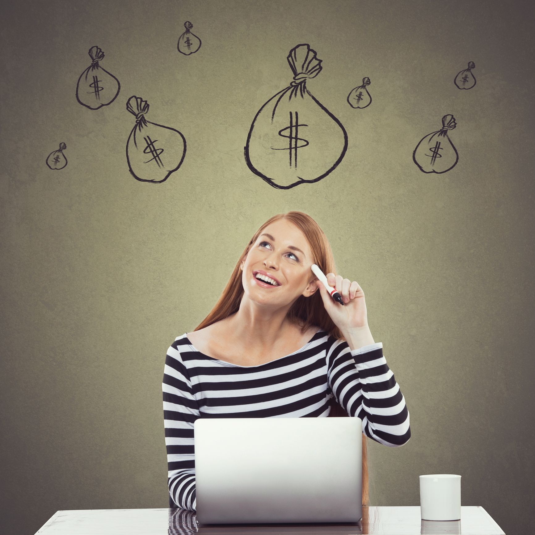 A woman is sitting at a desk with a laptop and looking up at drawings of money bags.